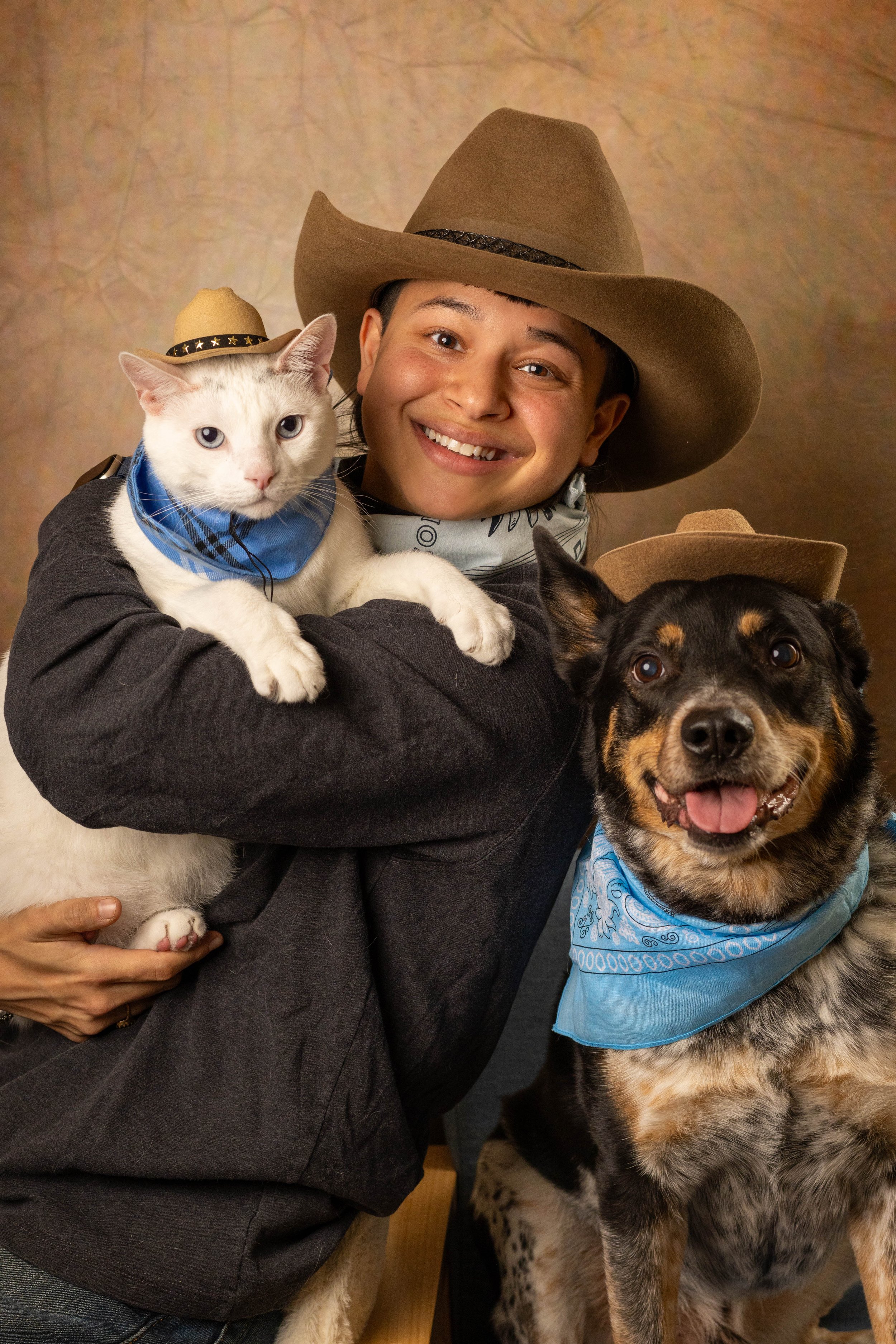 A person wearing a cowboy hat and a bandana around their neck, smiling and holding a white cat and a dog, both wearing small cowboy hats and bandanas.