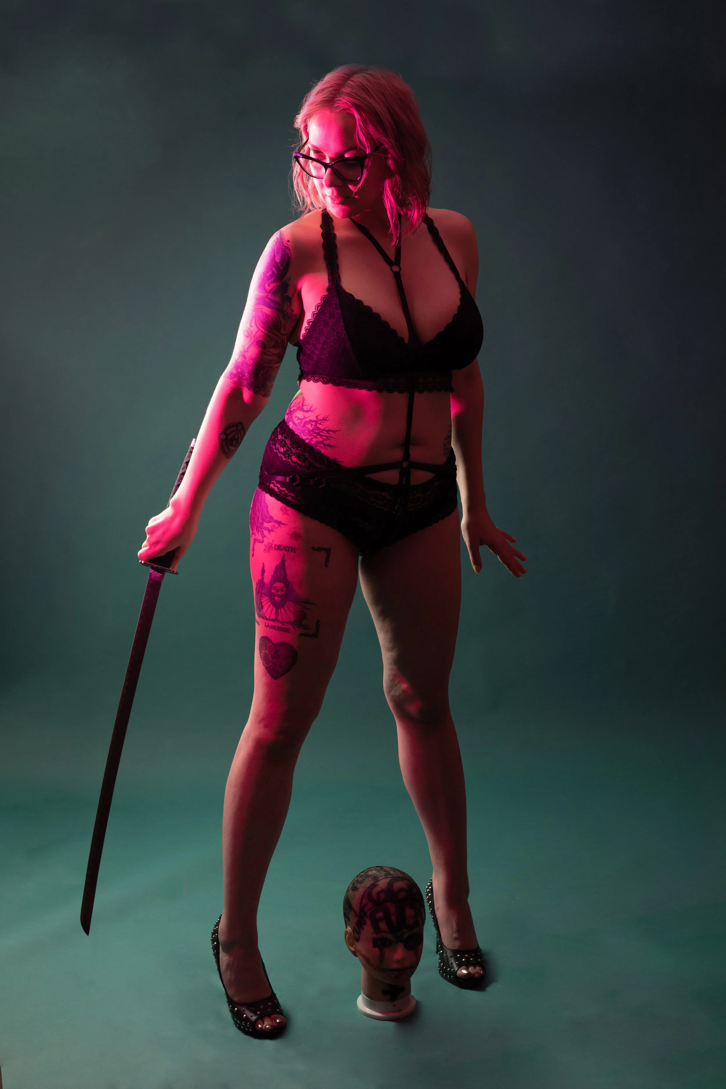 A woman with pink hair and glasses wearing black lingerie, holding a sword, standing on a green surface with a mannequin head at her feet.