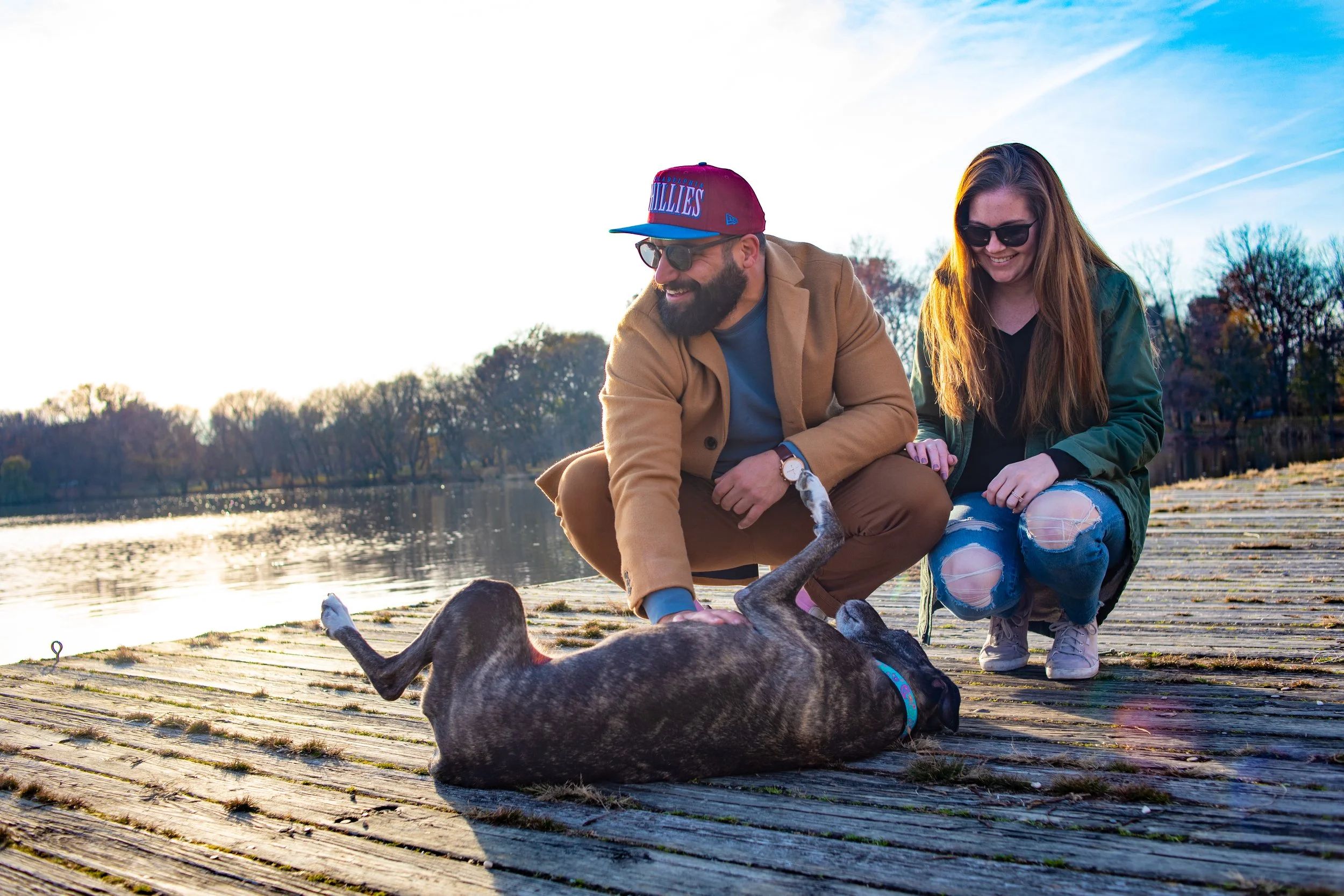 A man and woman enjoy playing with a dog on a wooden dock by a lake at sunset, with trees in the background and the sky showing a mix of blue and orange hues.