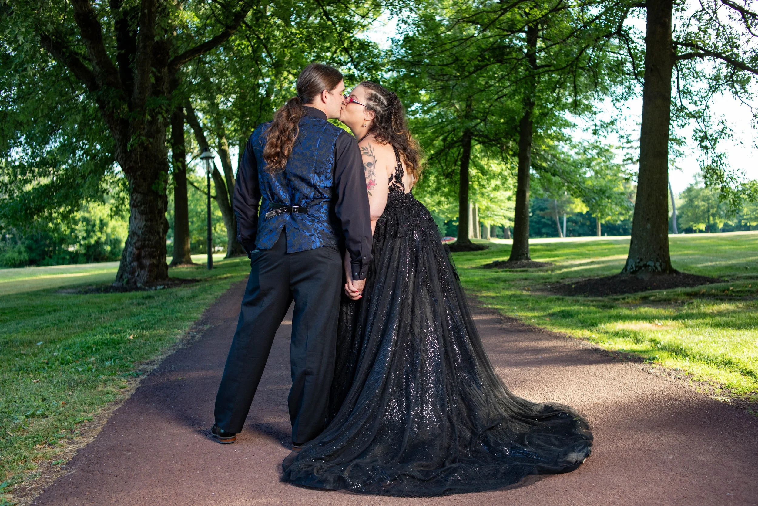 A couple sharing a kiss, holding hands, outdoors on a tree-lined path, one wearing a black gown and the other in a blue vest and black pants.