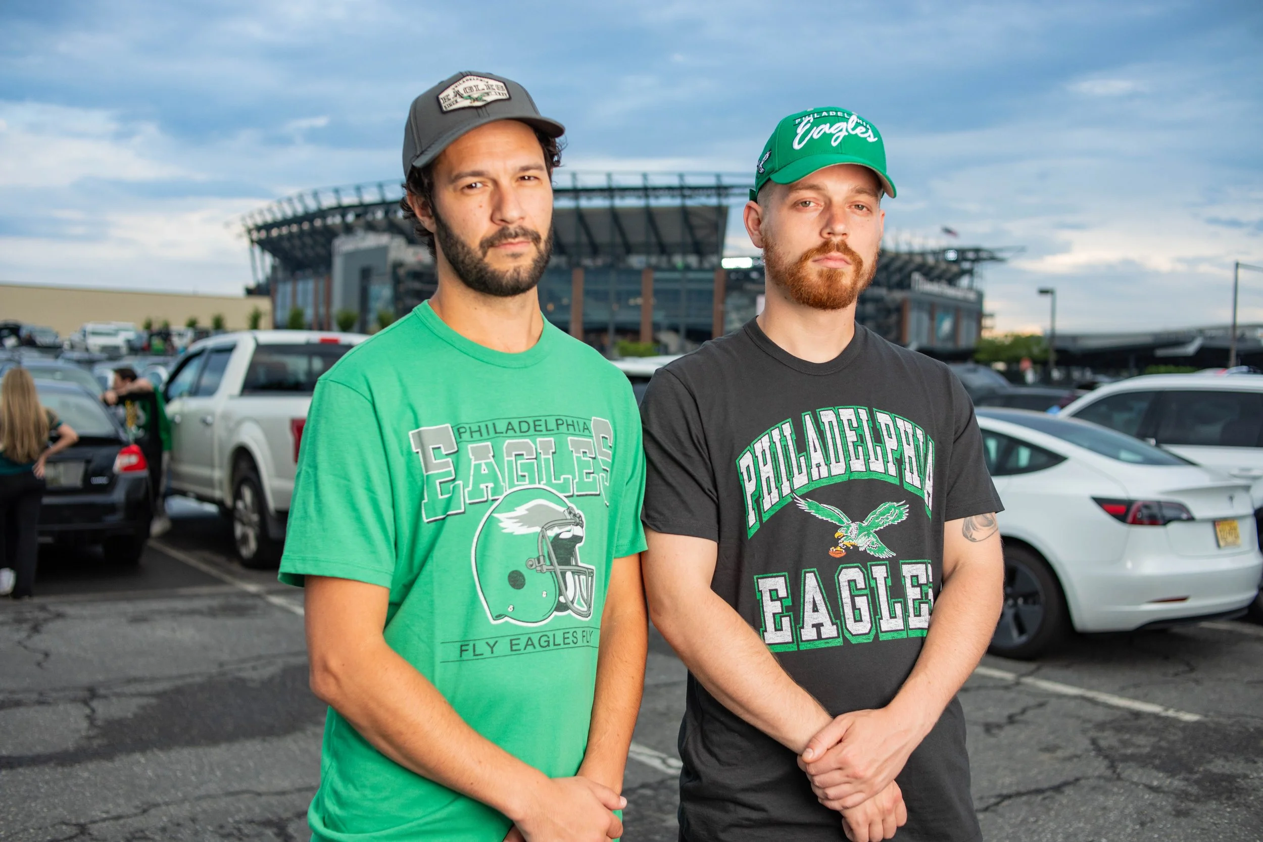 Two men standing in a parking lot with a stadium in the background, both wearing Philadelphia Eagles shirts and caps.