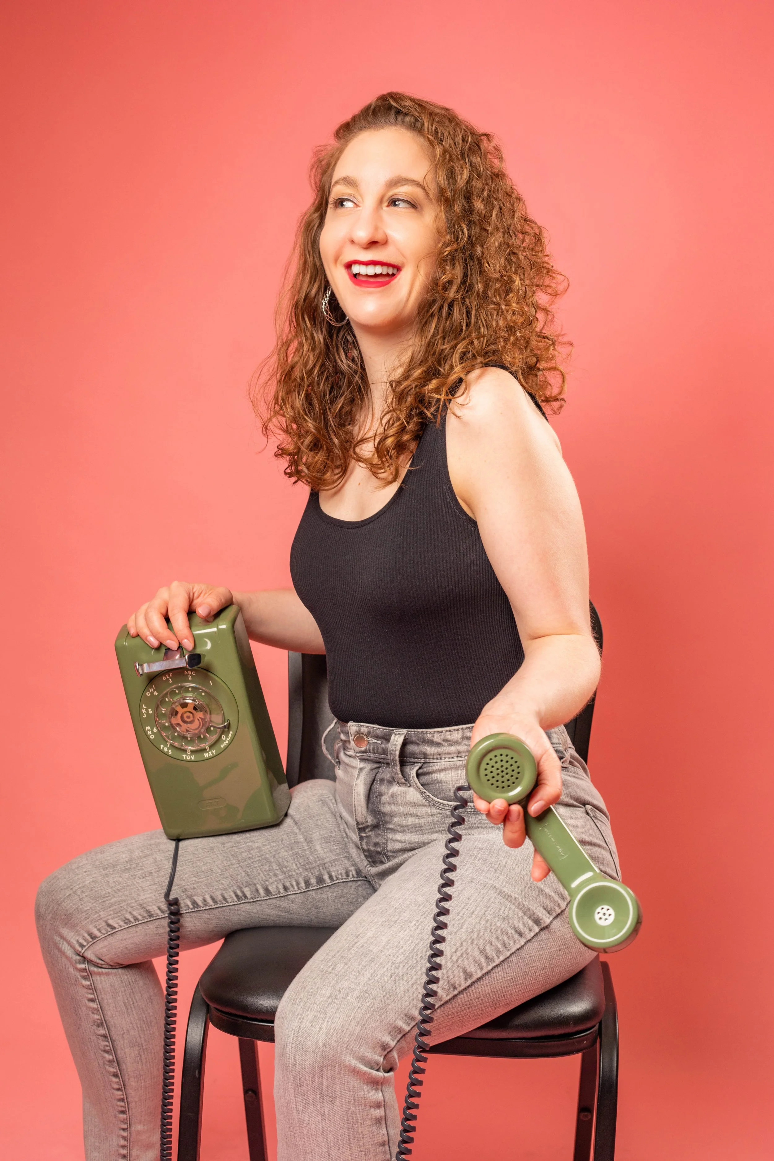 A woman with curly hair and a black tank top sitting on a black chair, holding a vintage green rotary phone and smiling, against a pink background.