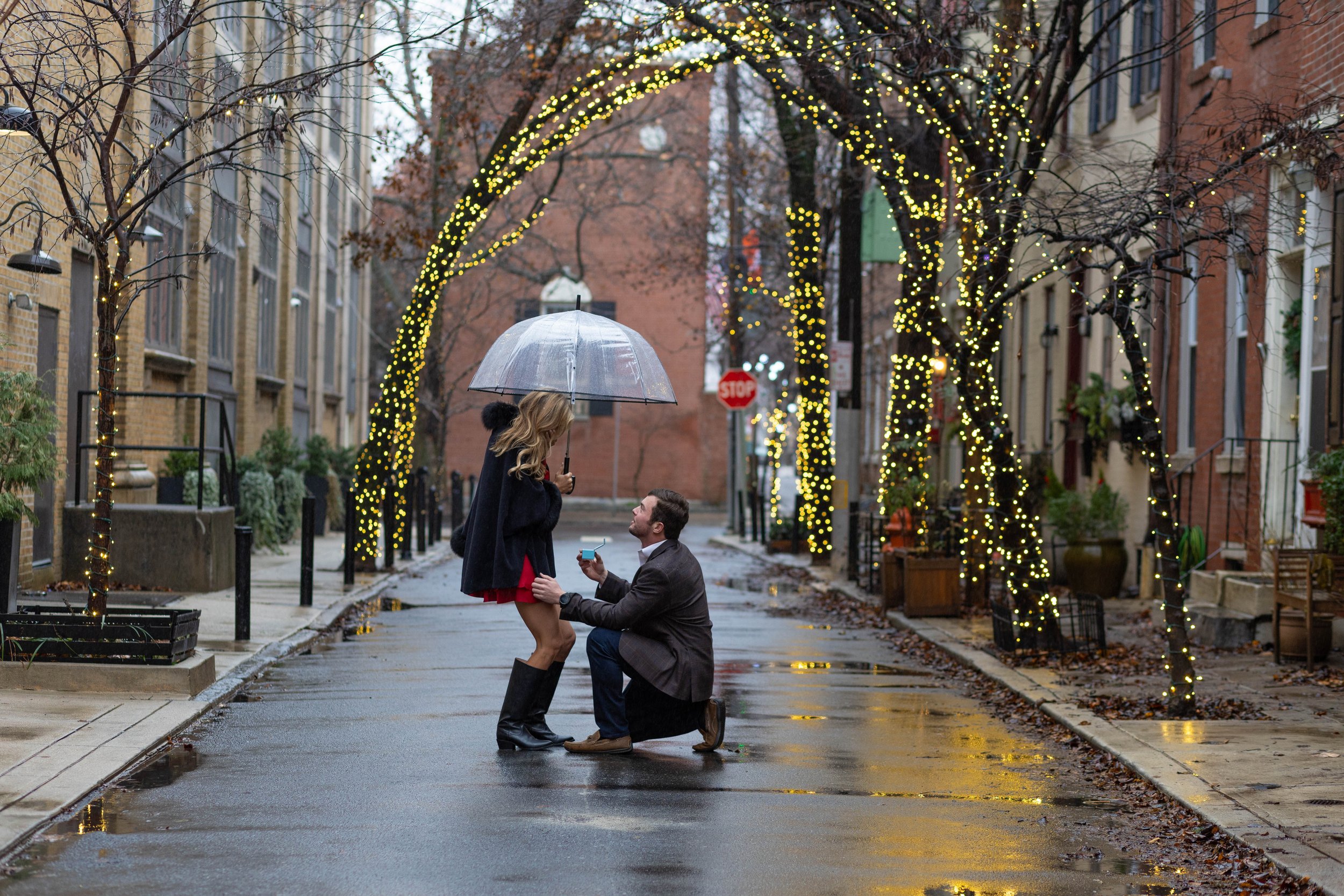 A man proposing to a woman on a rainy street decorated with string lights. The woman holds an umbrella, and the man is on one knee holding a ring box. The scene is festive and romantic.