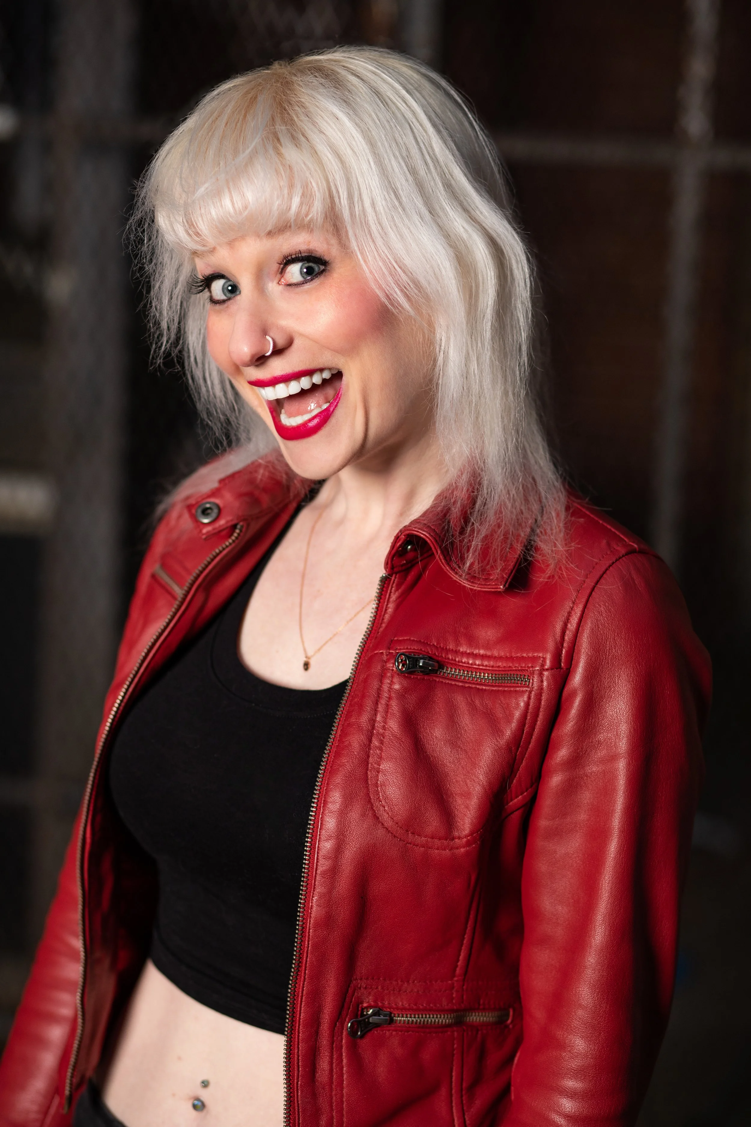 A woman with platinum blonde hair, red lipstick, and a nose piercing, smiling and looking at the camera, wearing a red leather jacket over a black crop top, with a blurred industrial background.
