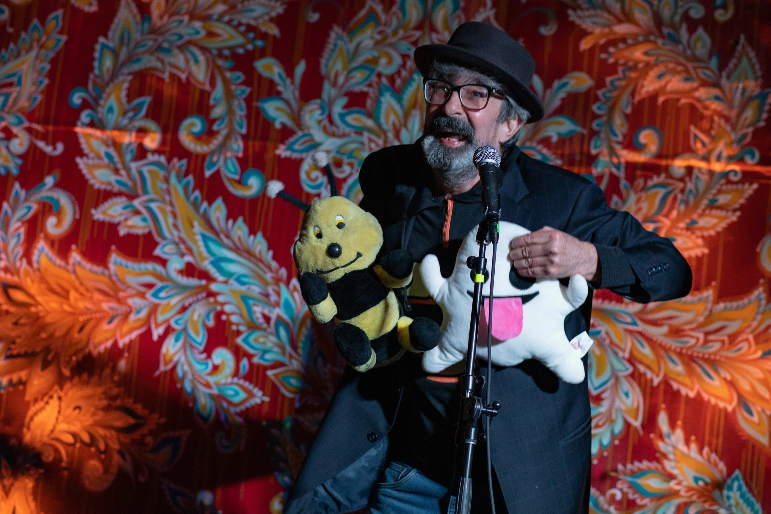 A man with a beard and glasses, wearing a black hat, black jacket, and orange shirt, is speaking into a microphone. He is holding and wearing stuffed toys of Woodstock and Snoopy from Peanuts. The background features a colorful, patterned fabric with