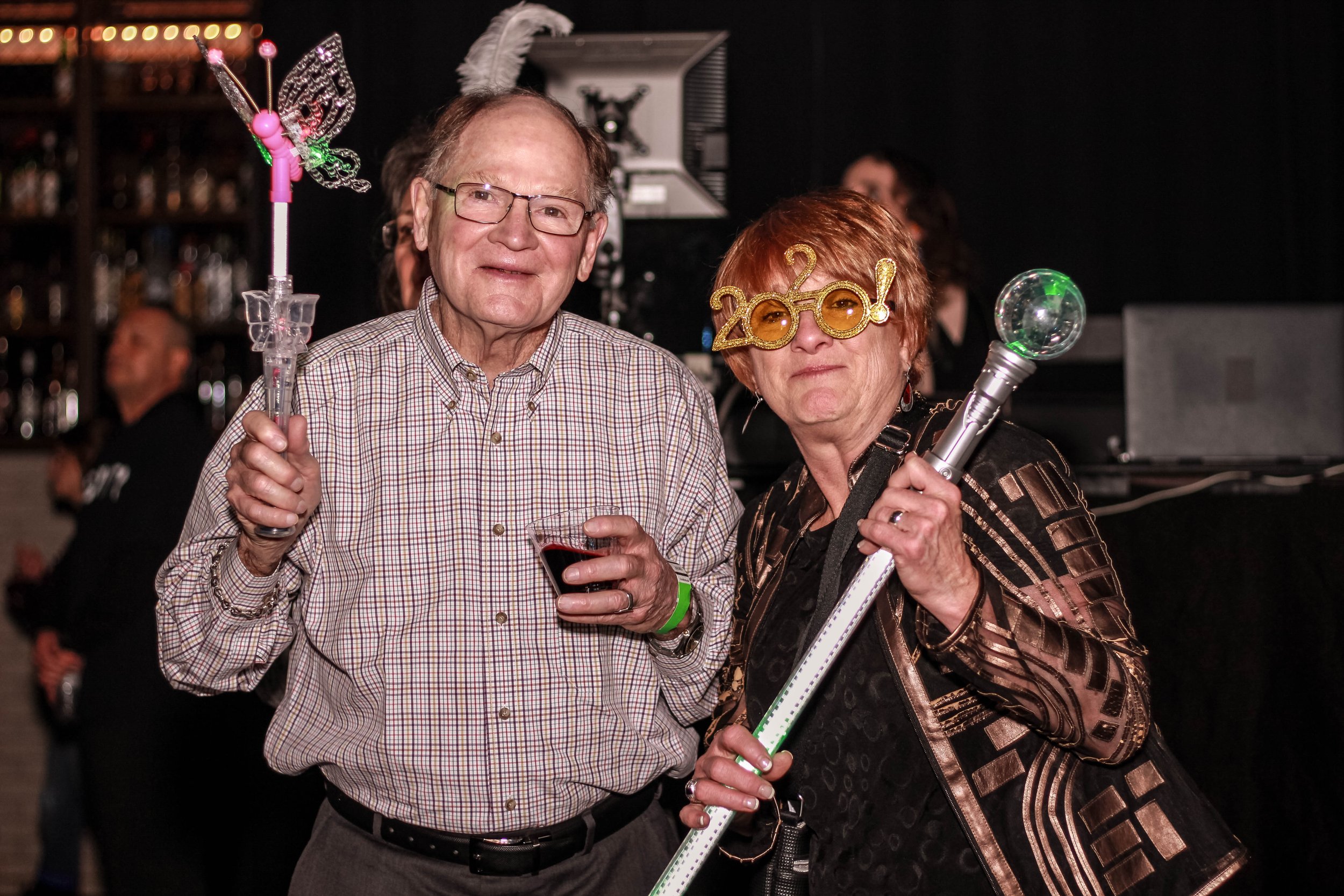 Older man and woman celebrating New Year's Eve at a party, holding glow sticks, with festive glasses and accessories.