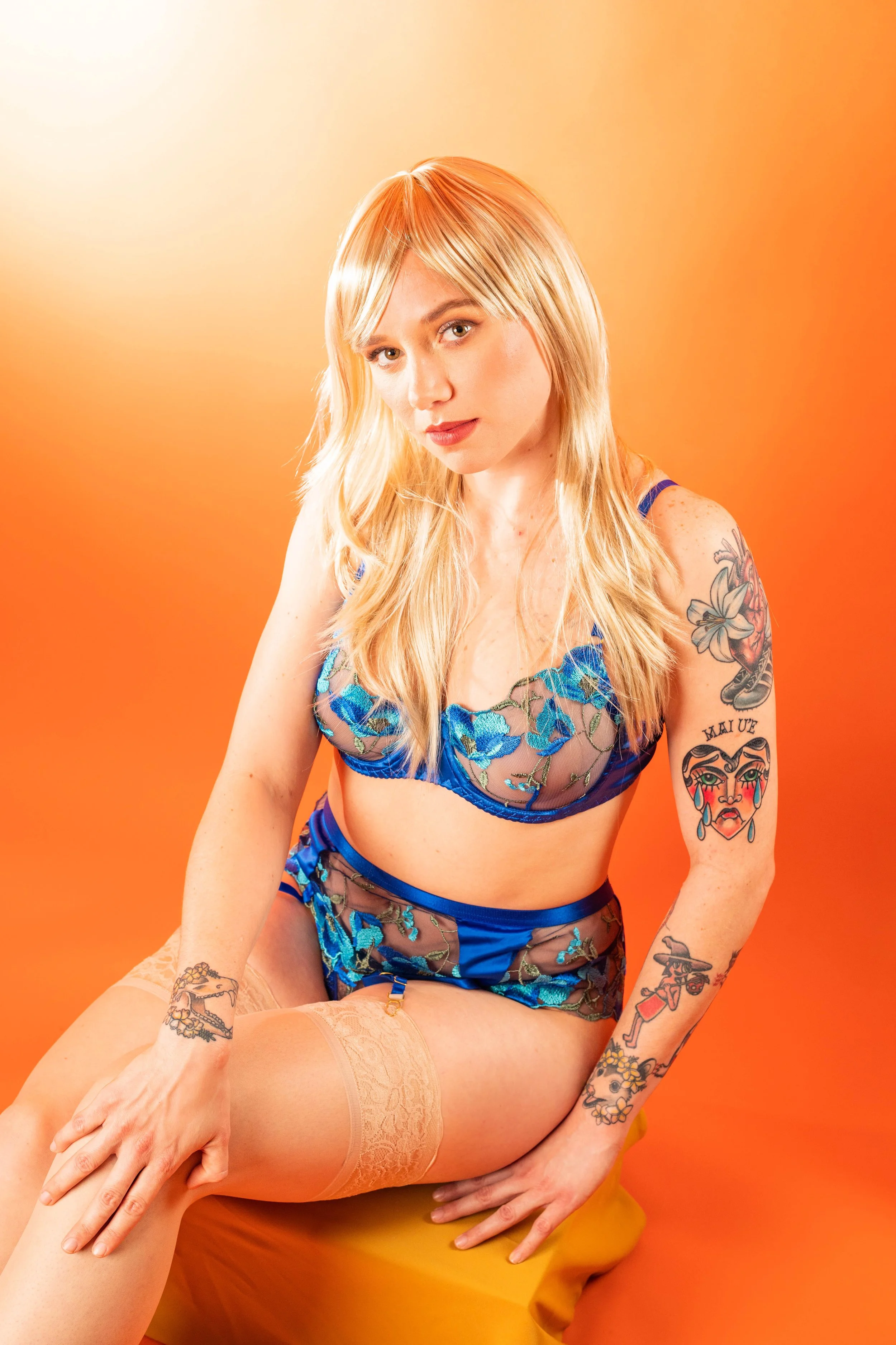 A woman with long blonde hair wearing blue lace lingerie with floral embroidery, sitting on a yellow surface against an orange background.