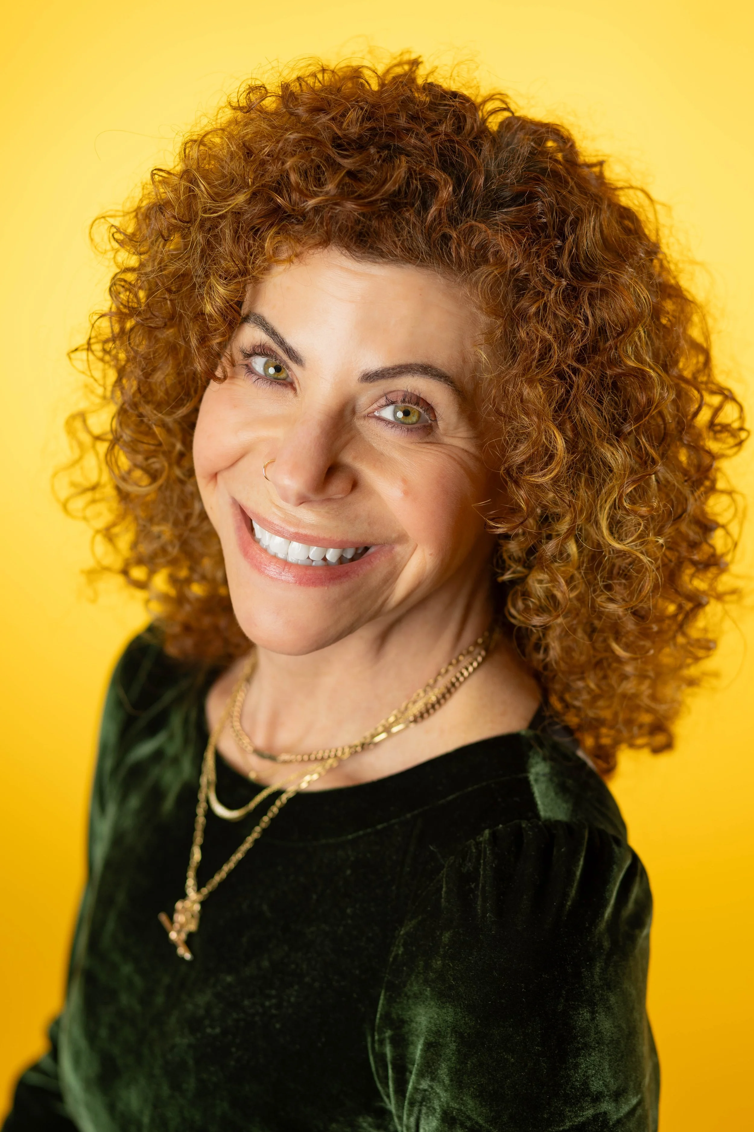 A woman with curly red hair smiling, wearing a black velvet top and gold jewelry, standing against a bright yellow background.