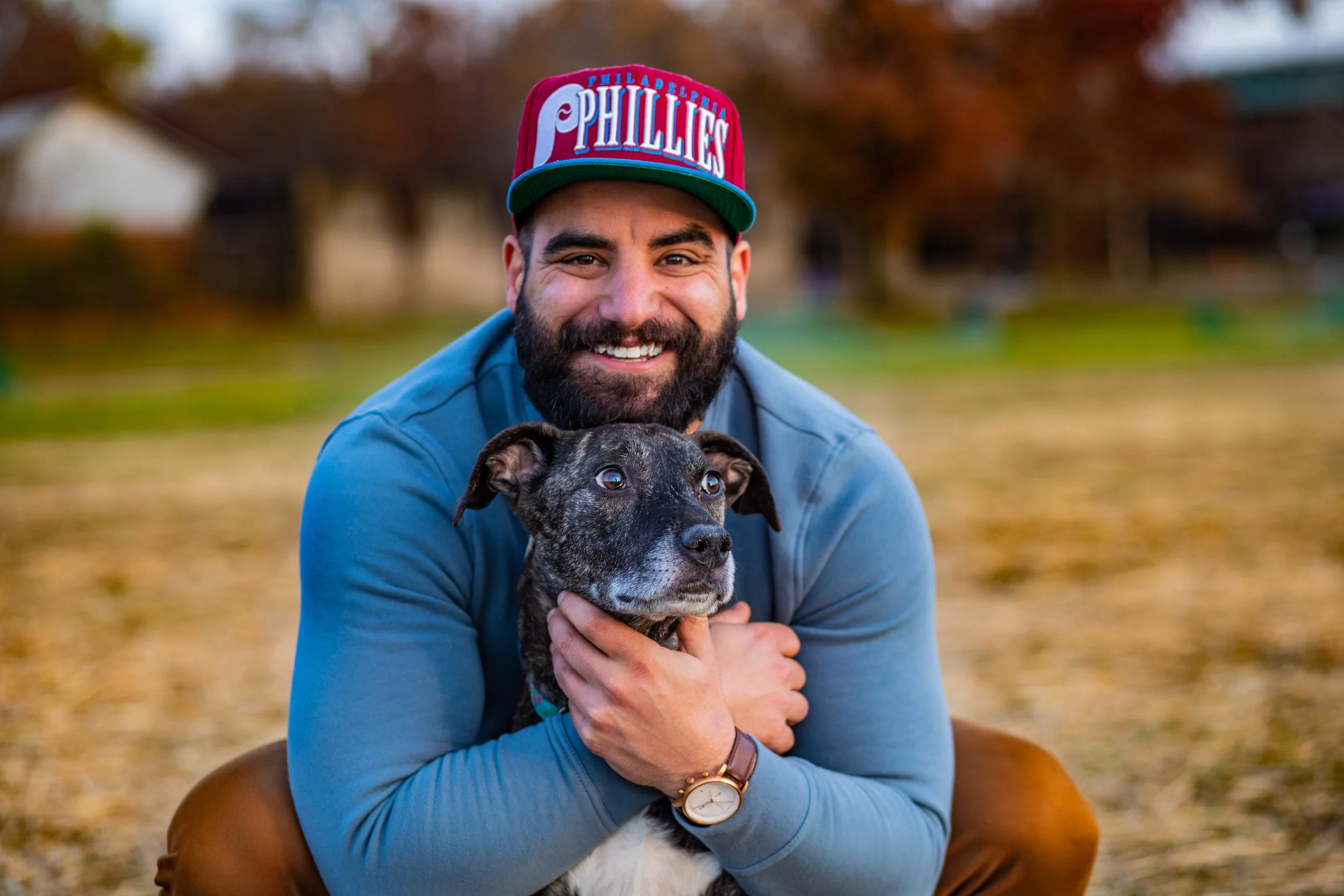 A smiling man with a beard wearing a red and green Philadelphia Phillies baseball cap, holding a brindle and white mixed-breed dog outdoors in a park during fall.