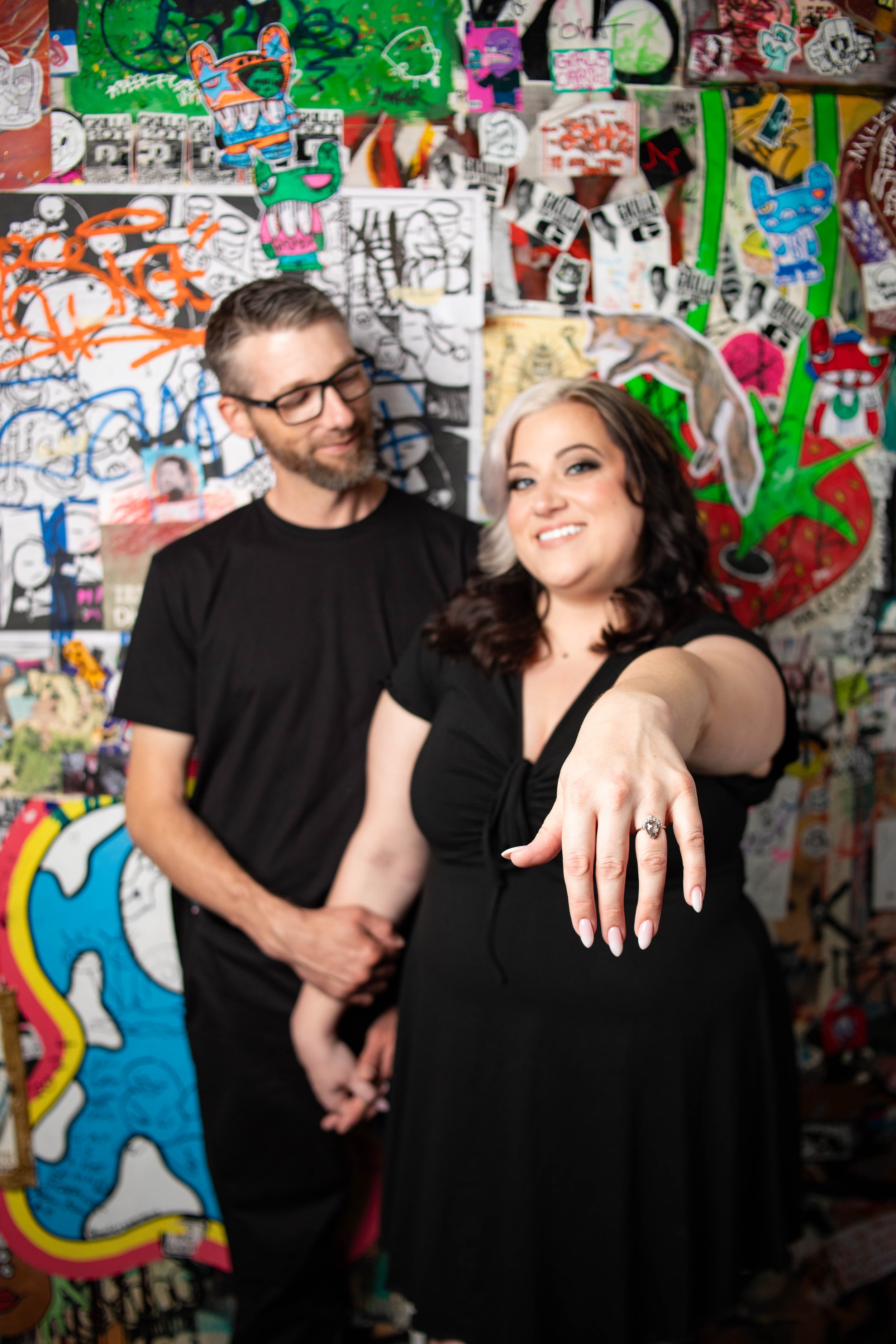 A woman showing her engagement ring while a man stands behind her, both smiling, in front of a colorful graffiti wall.