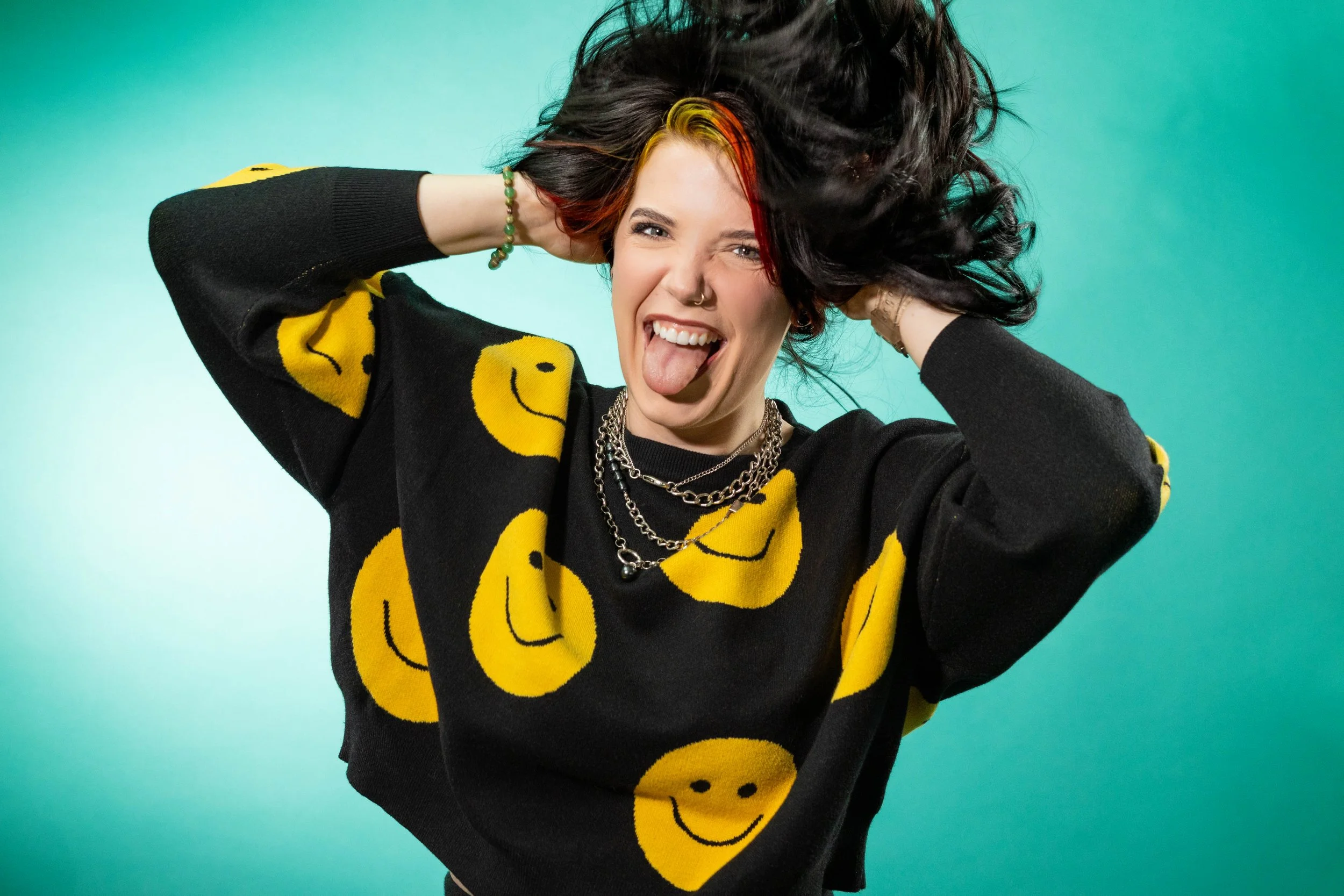 A young woman with black and red hair, sticking out her tongue, and making a silly face. She is wearing a black sweater with yellow smiley faces, layered necklaces, a nose ring, and bracelets. She is standing against a green background with her arms 