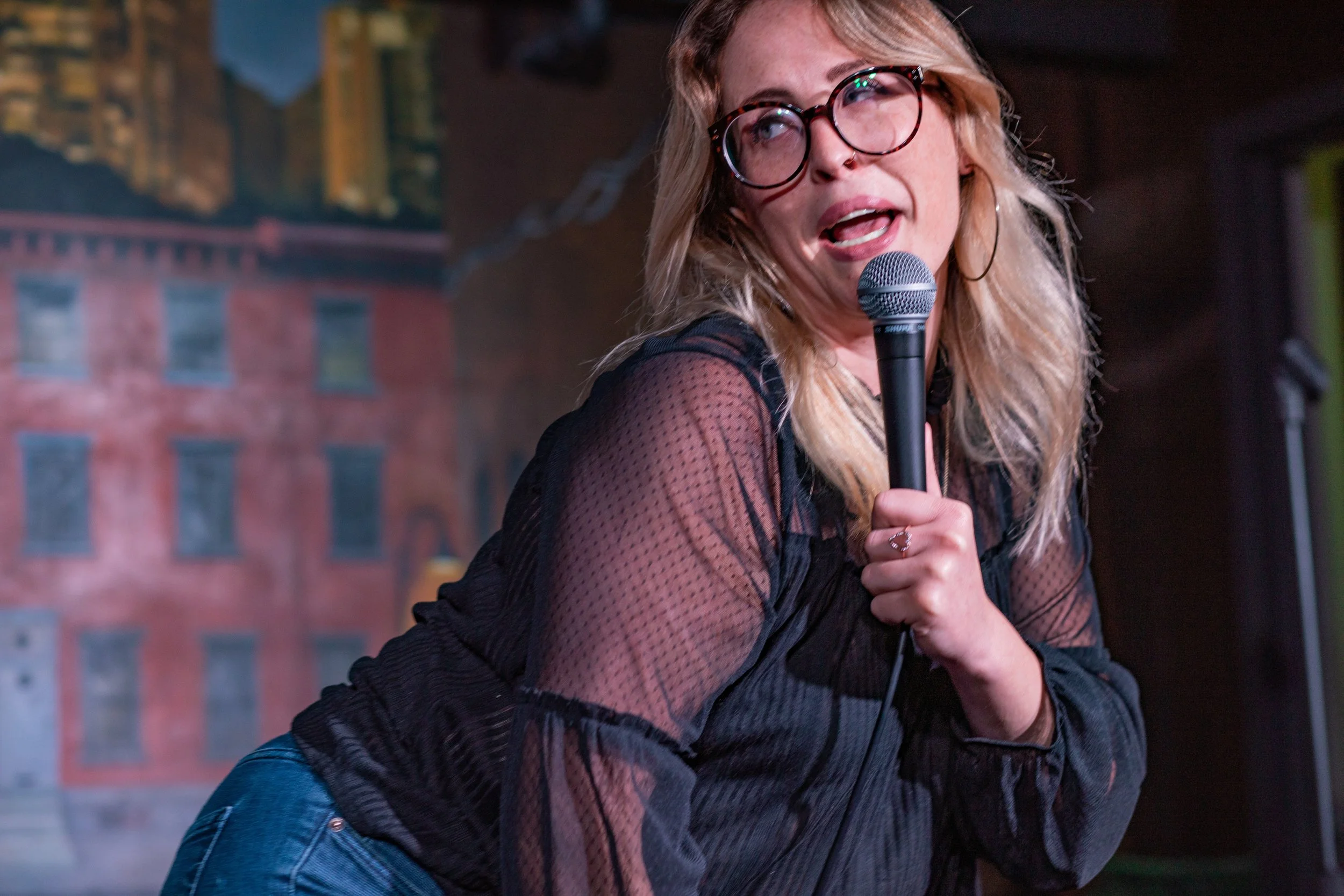 A woman with blonde hair, glasses, and hoop earrings performing on stage with a microphone, wearing a black sheer blouse, with a backdrop of a cityscape painting.