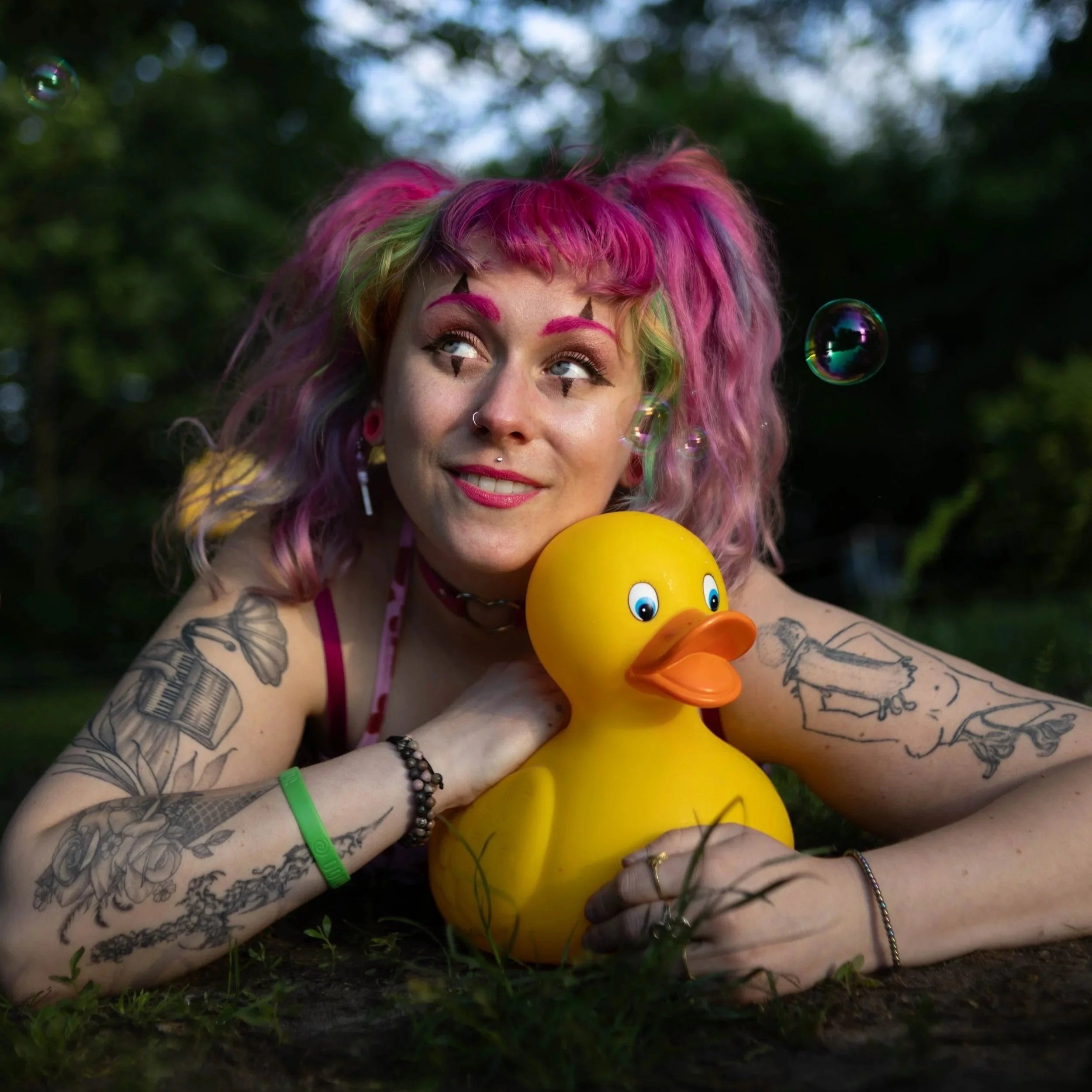 A woman with pink and green hair, tattoos, and colorful makeup is lying on the grass outdoors, holding a yellow rubber duck and surrounded by floating soap bubbles.