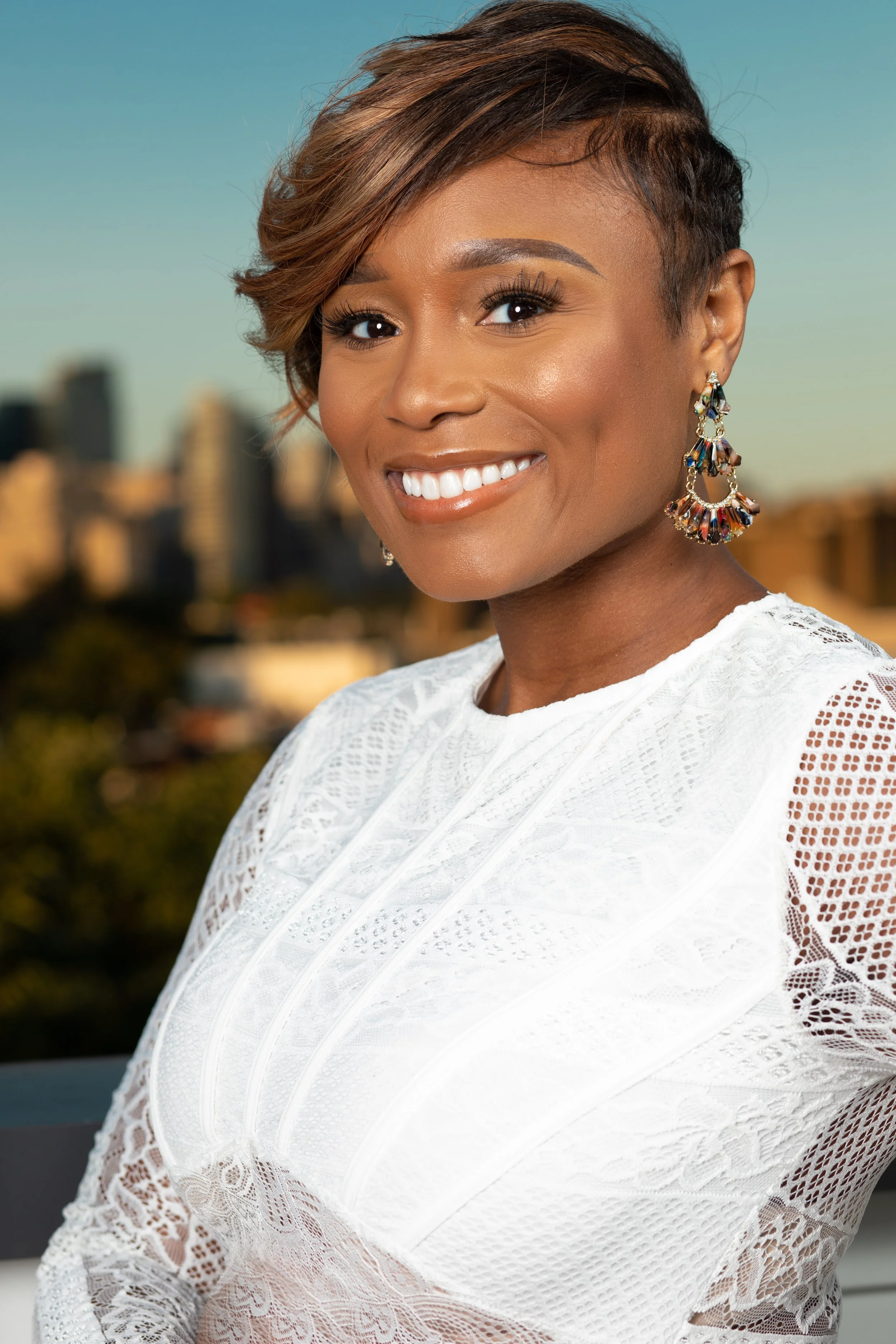 A woman smiling outdoors with blurred city skyline in the background, wearing white lace top and colorful earrings.