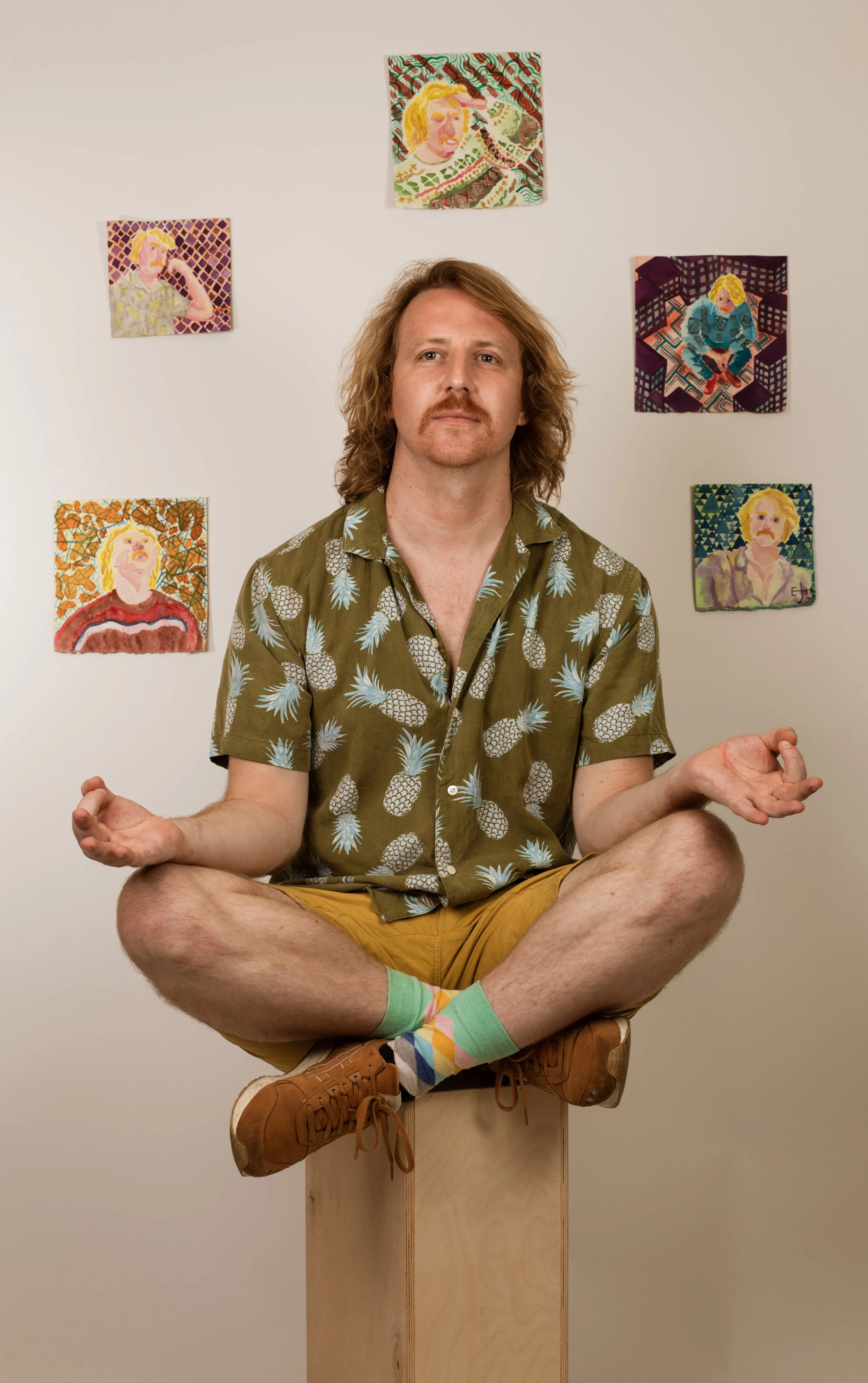 A man with long hair, a mustache, and a goatee sitting cross-legged on a wooden block, with arms outstretched and palms up, wearing a pineapple patterned shirt, tan shorts, and colorful socks. Behind him are six colorful portrait paintings on a white