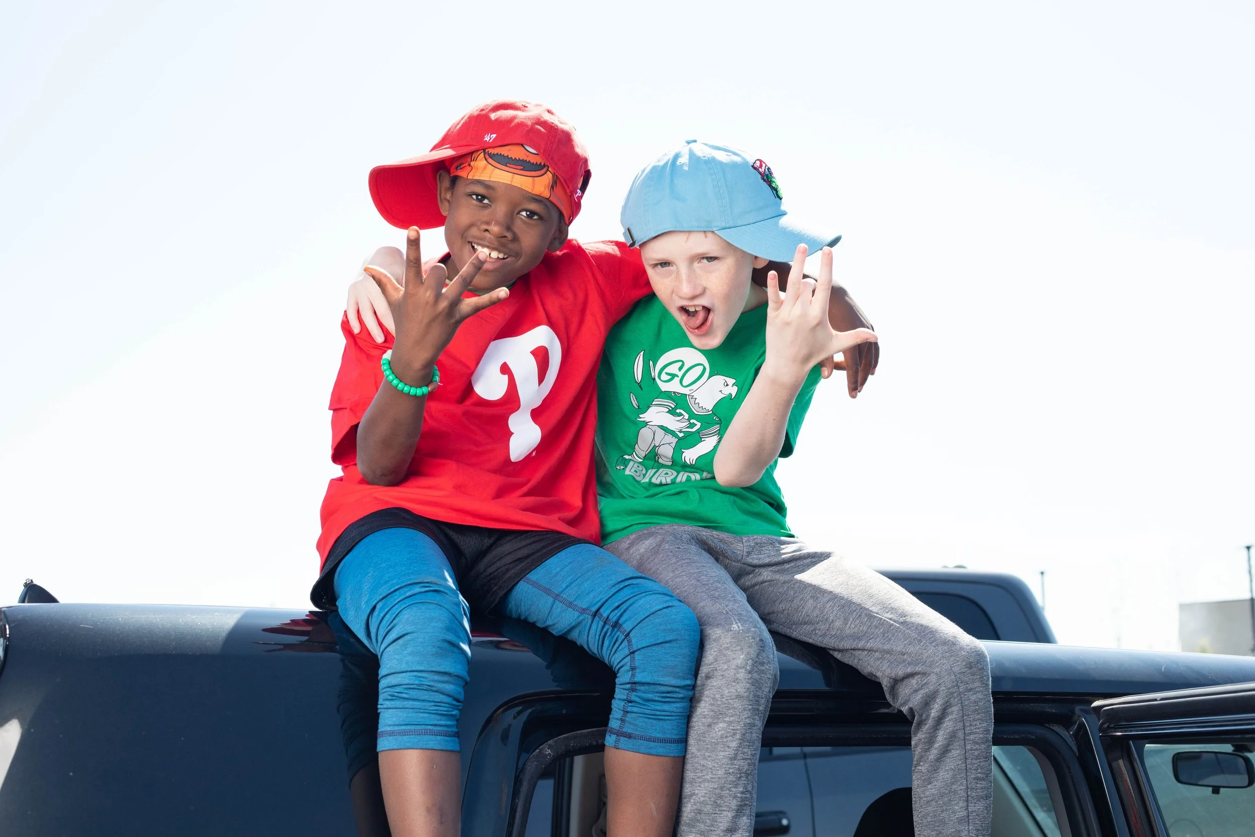 Two boys sitting on the roof of a vehicle, smiling and making peace signs. One wearing a red cap backwards, a red shirt, and blue striped pants. The other wearing a blue cap, a green shirt, and gray pants.
