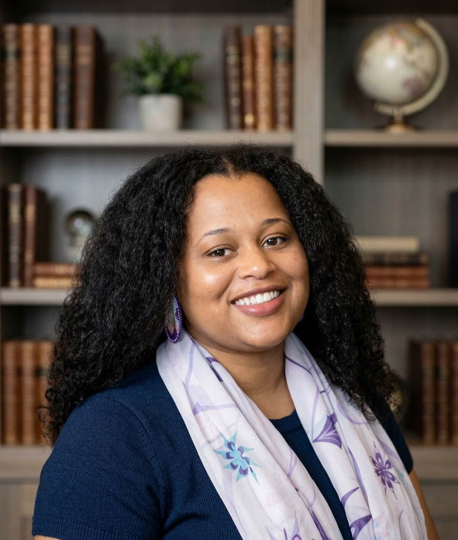 A woman with curly black hair smiling at the camera, wearing a dark blue top and a light-colored floral scarf, in front of a bookshelf with books and a globe.