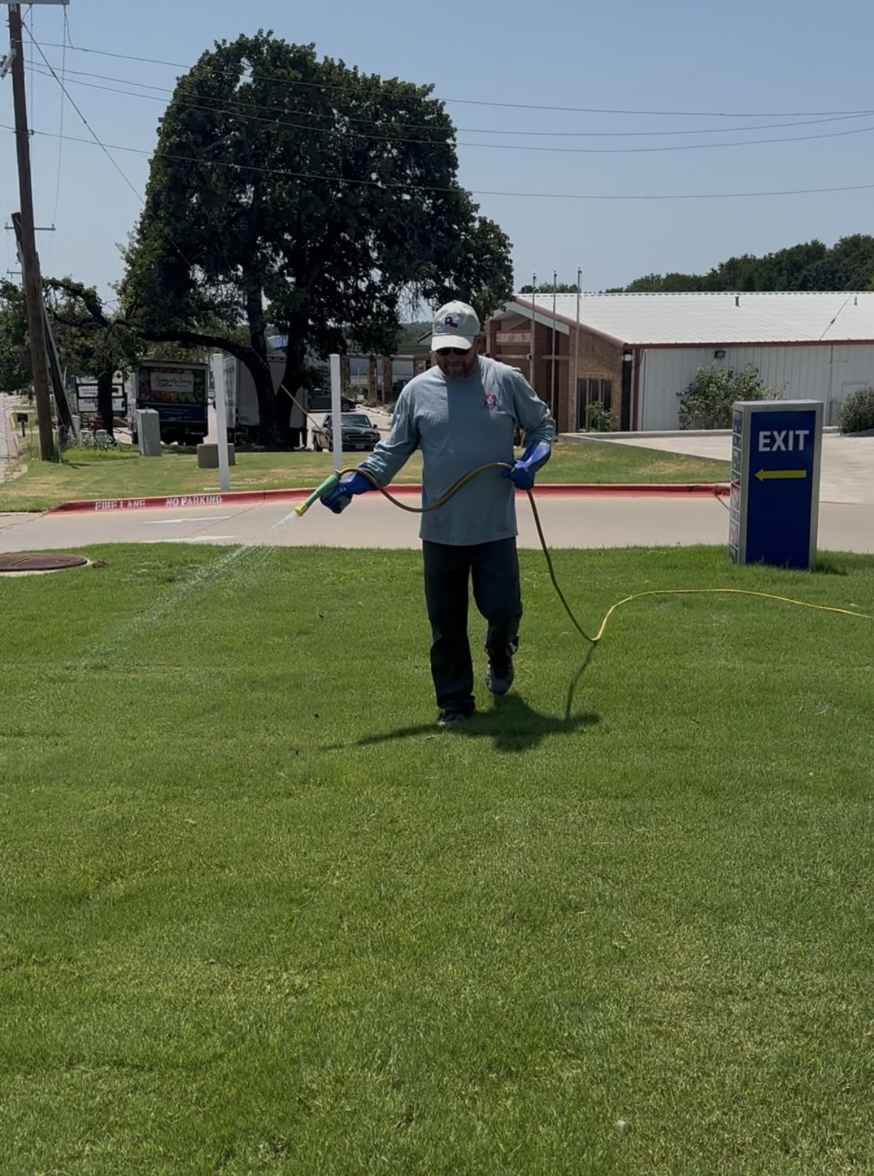 A man in a gray uniform and white cap spray-paints grass with a garden sprayer on a sunny day.