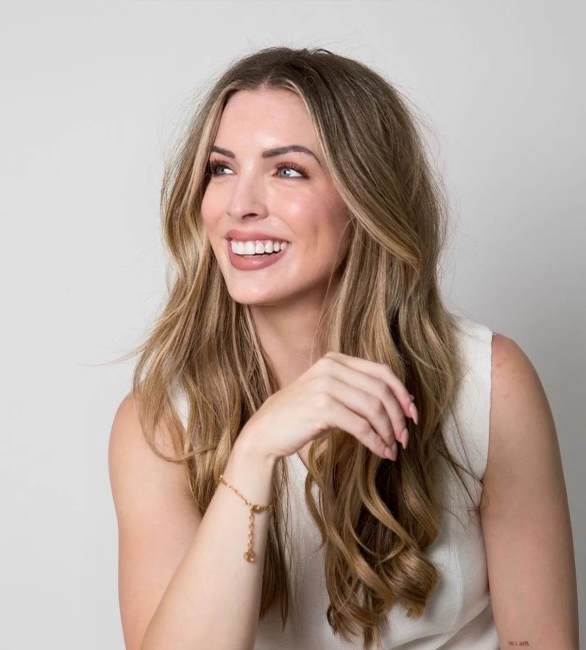 Smiling woman with long wavy brown hair wearing a sleeveless white top against a light gray background