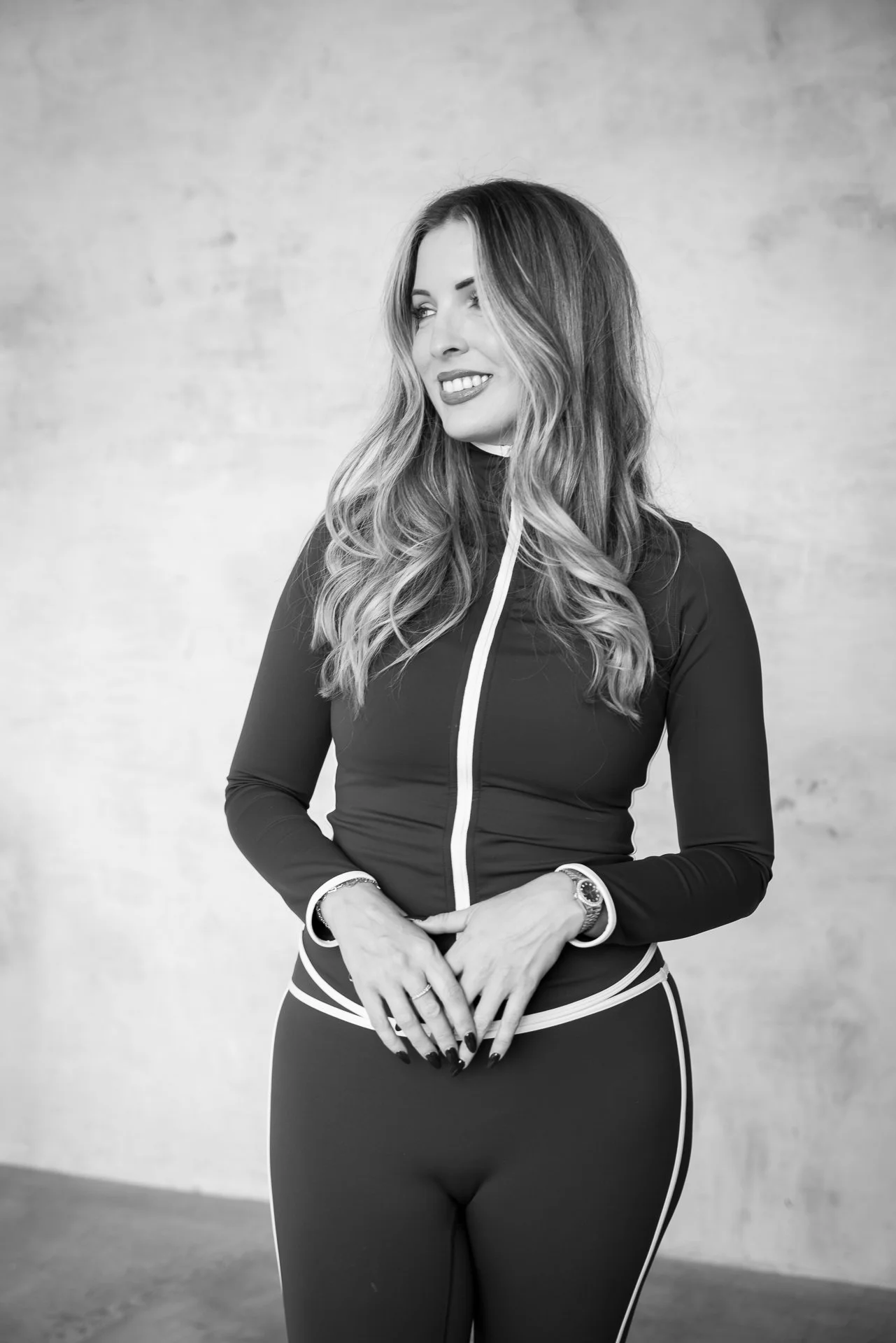 Black-and-white portrait of a smiling woman with long wavy hair wearing a fitted zip-up top and leggings.
