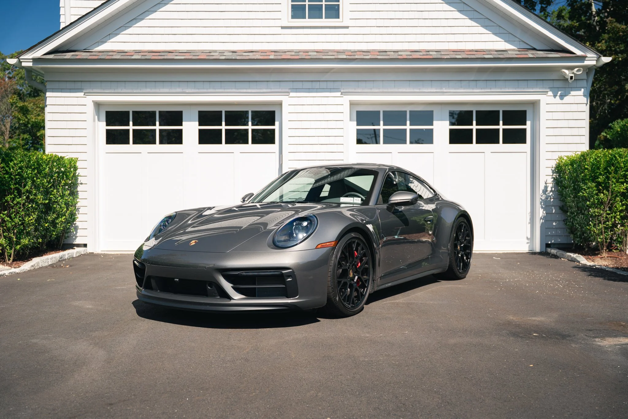 Gray Porsche sports car parked in front of a white garage with double doors, green bushes on each side, and a paved driveway.