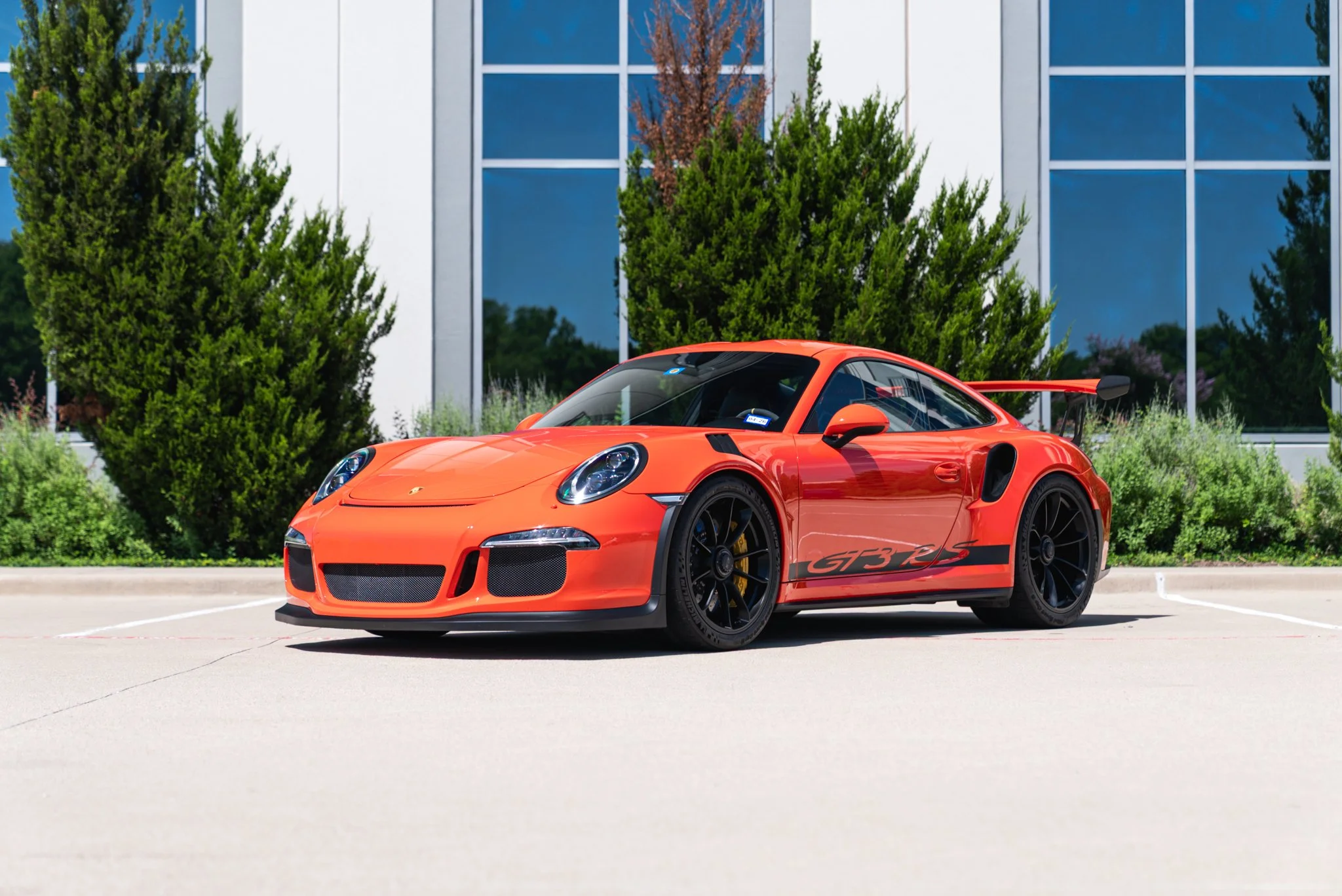 Orange Porsche 911 GT3 RS sports car parked in front of a modern building with glass windows and greenery.