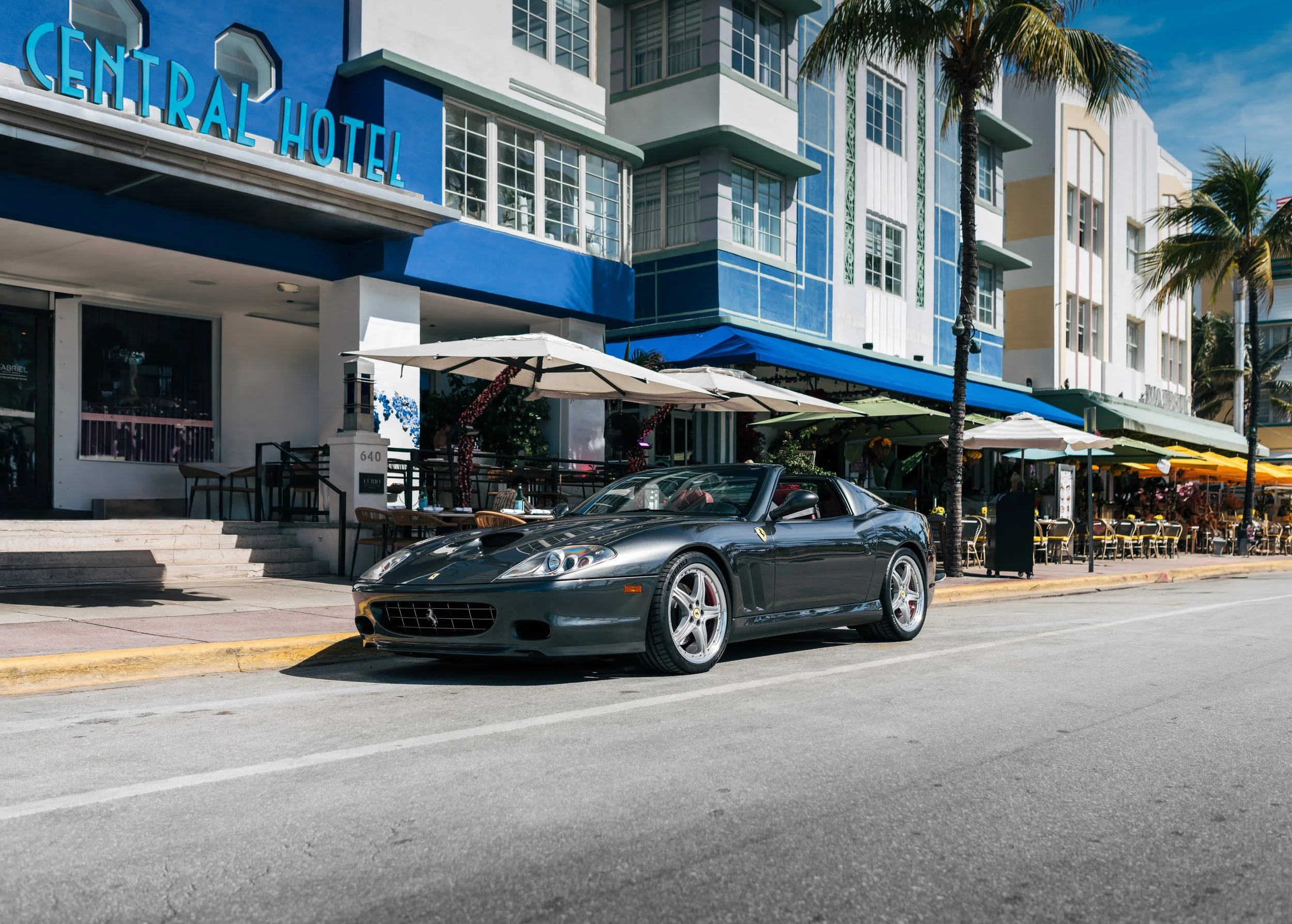 Black Ferrari sports car parked on a city street in front of a restaurant with outdoor seating and umbrellas, with colorful buildings and palm trees in the background under a clear blue sky.