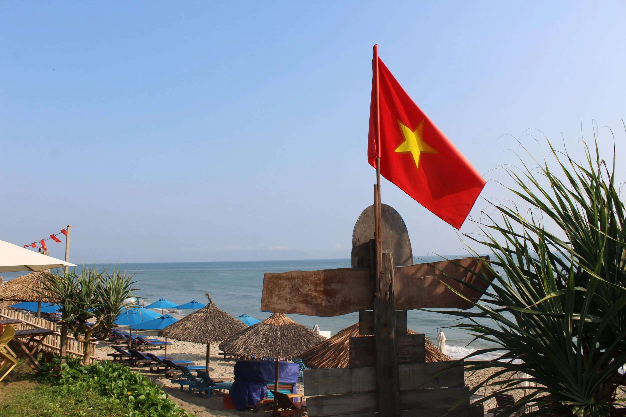 Vietnamese flag flying on a beach with thatched umbrellas, lounge chairs, palm trees, and the ocean in the background.