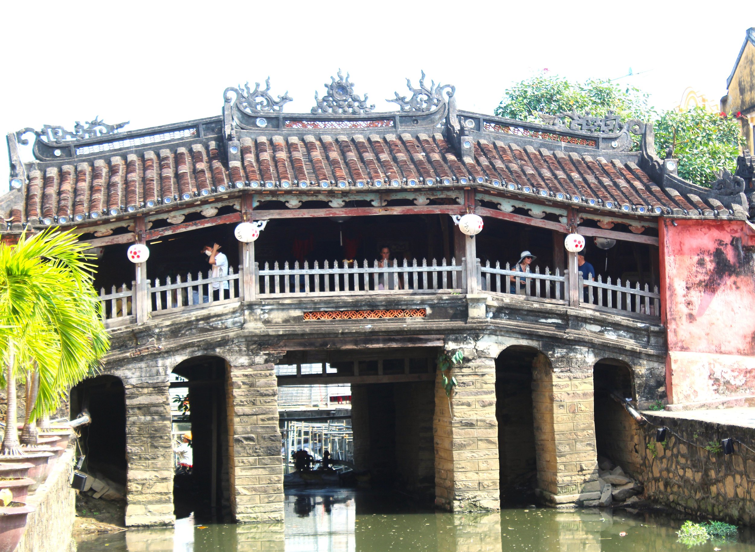 Hoi An's most famous bridge. A traditional Vietnamese two-story wooden house with ornate roof decorations and lanterns hanging, situated over water with stone pillars supporting the structure, surrounded by green plants and trees.