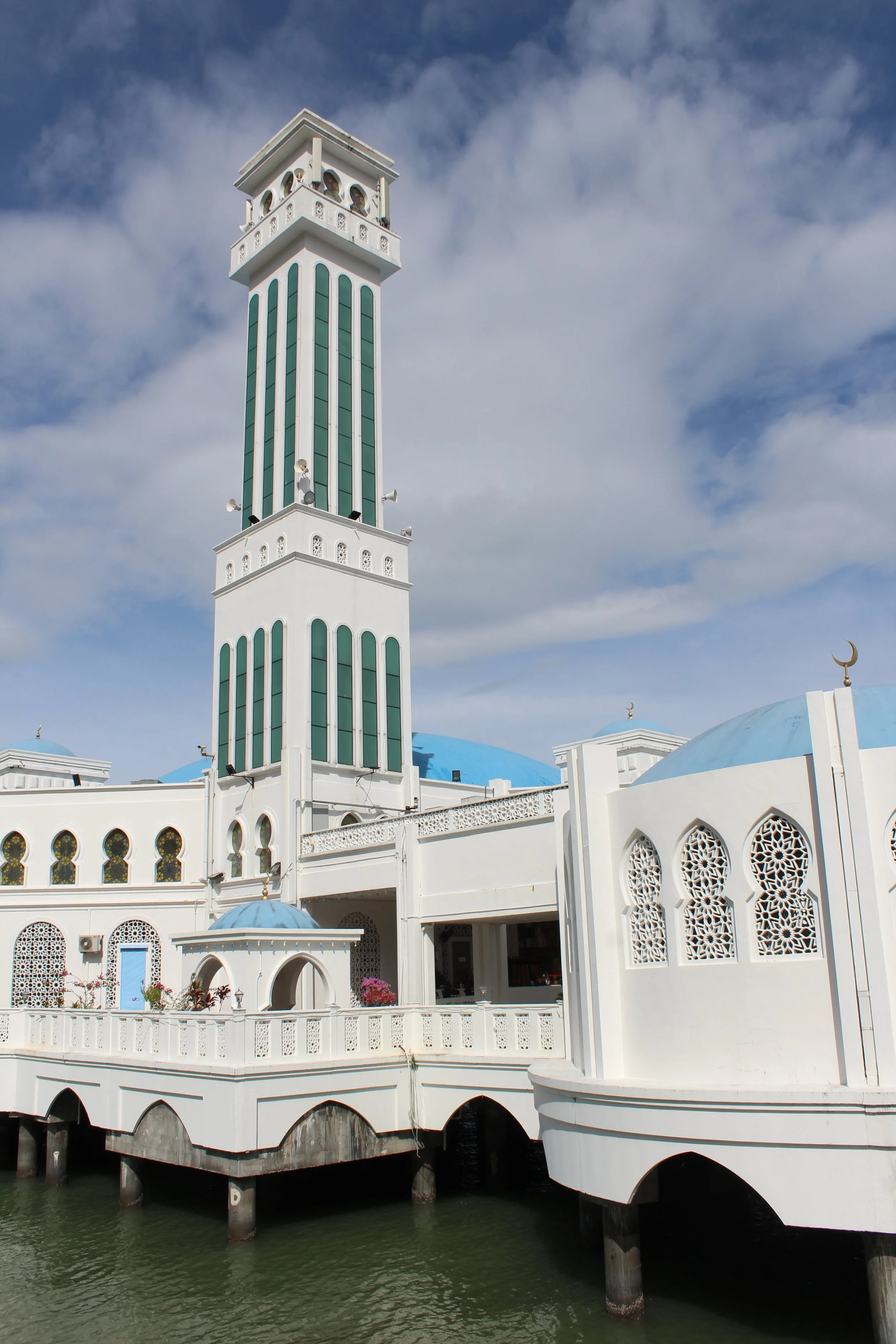 The Floating Mosque of Tanjung Bungah, Penang with blue domes and a tall minaret near a body of water against a partly cloudy sky.