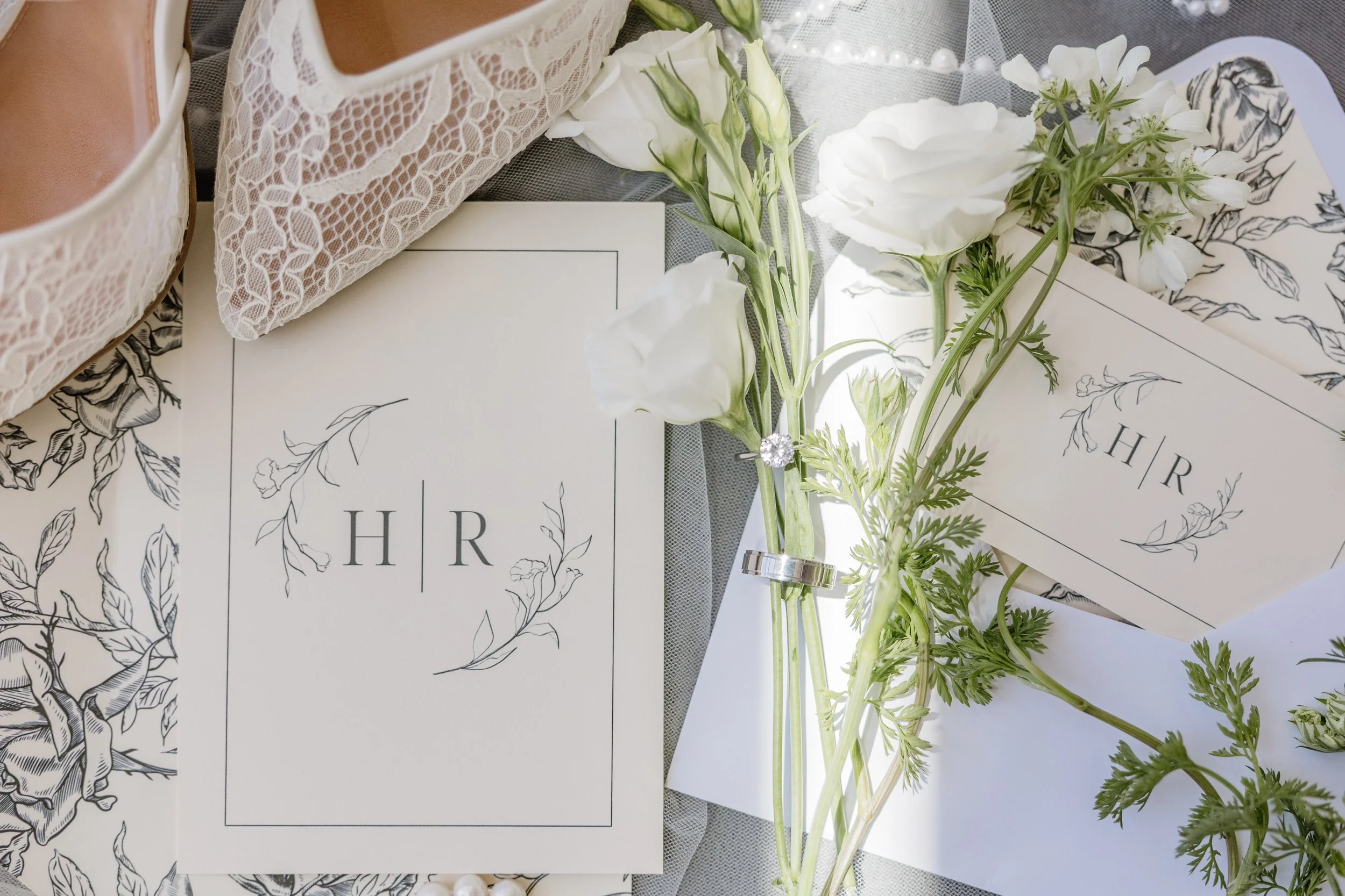 White flowers, black and white floral printed paper, wedding rings, pearl necklace, and wedding invitations with the initials 'HR'.
