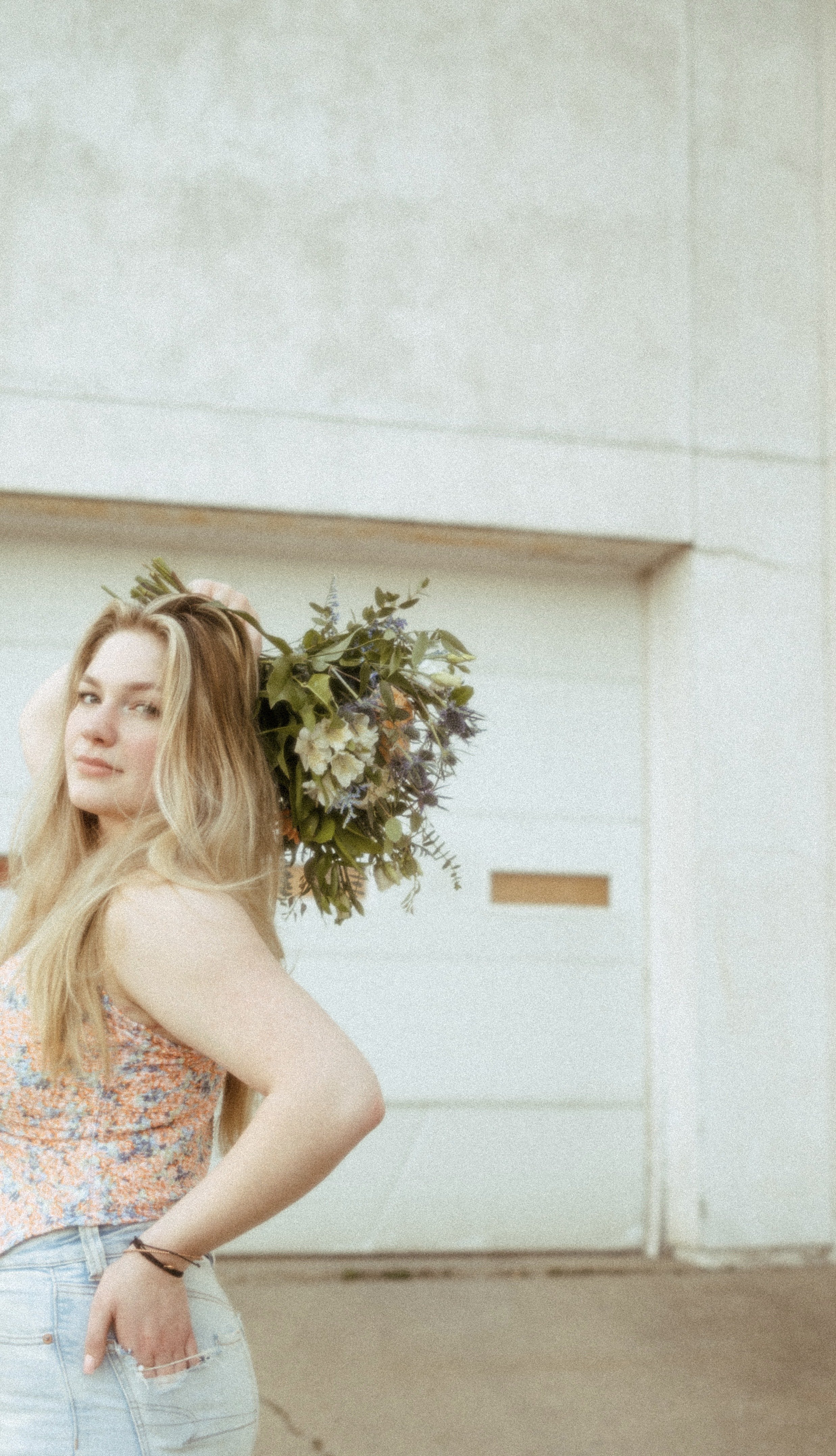 Woman with long blonde hair wearing a floral top and light jeans, holding a bouquet of flowers over her shoulder, standing outside near a white garage door.