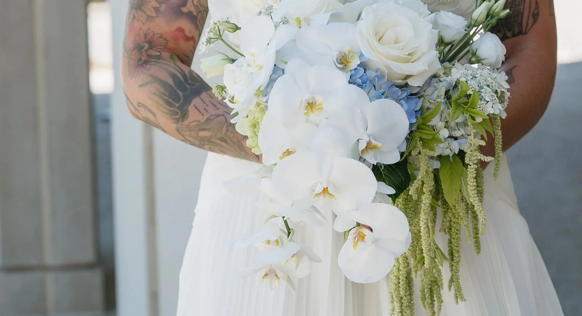 Close-up of a bride holding a cascading bouquet of white roses, orchids, and greenery, with tattooed arms visible.