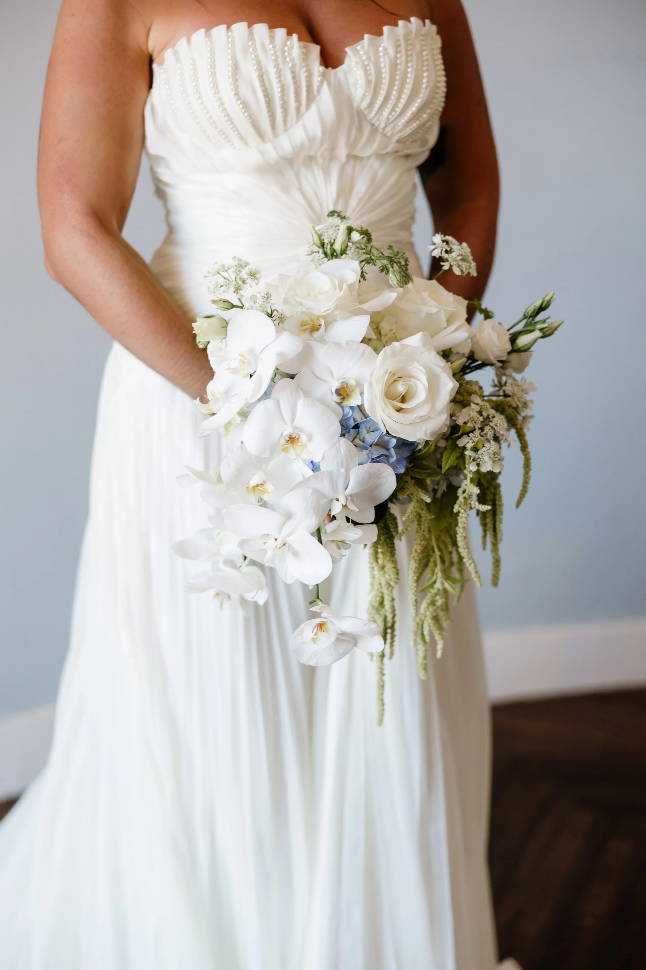 A woman in a white satin wedding dress holding a cascading bouquet of white roses and orchids.
