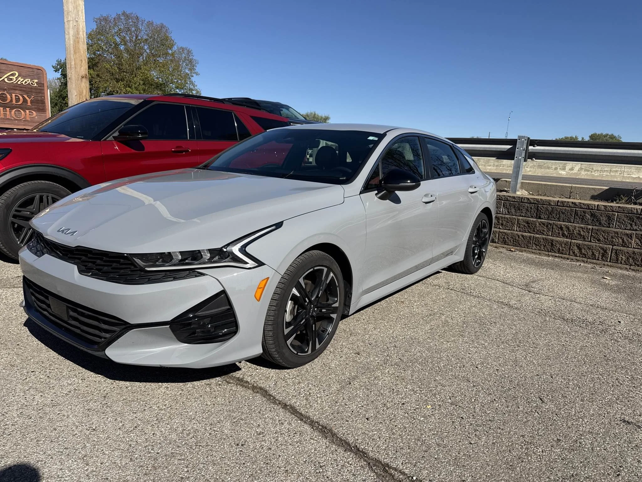 A silver Kia sedan parked next to a red vehicle in a parking lot with a brick wall and guardrail in the background, clear sky overhead.