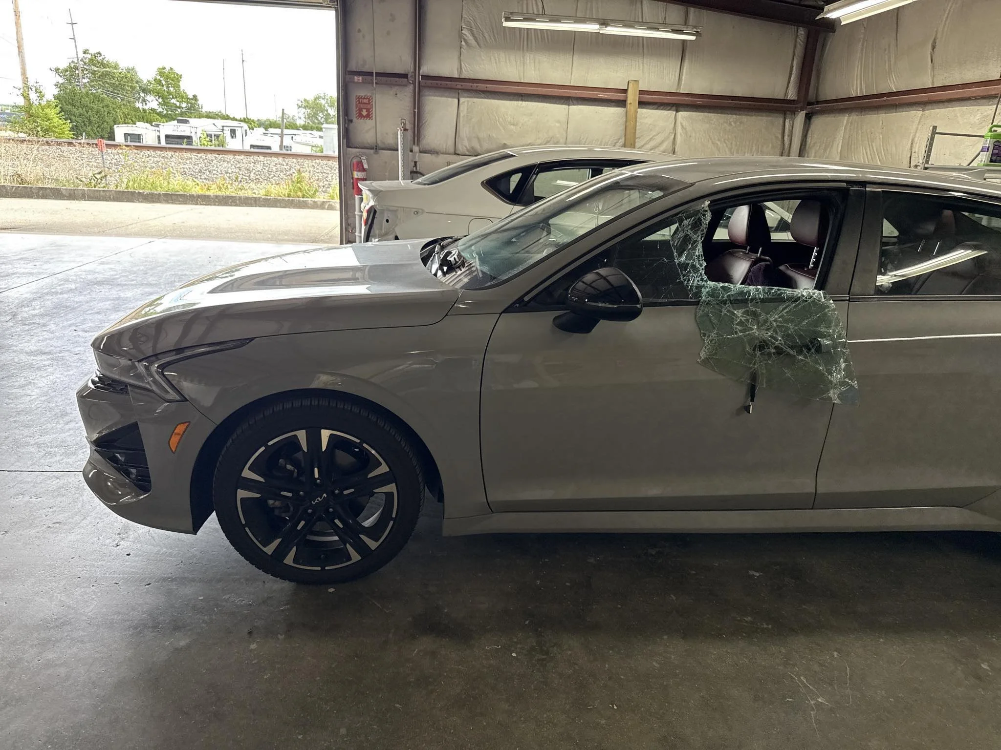 A silver car with a shattered driver side window and front end damage parked inside a garage, with a white car in the background.