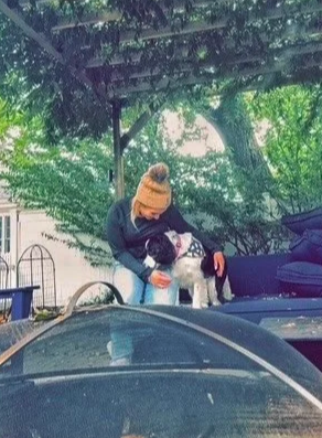 Person wearing a brown knit hat and dark clothing sitting outdoors, petting a small white dog under a shaded area with trees and a dark-colored couch.