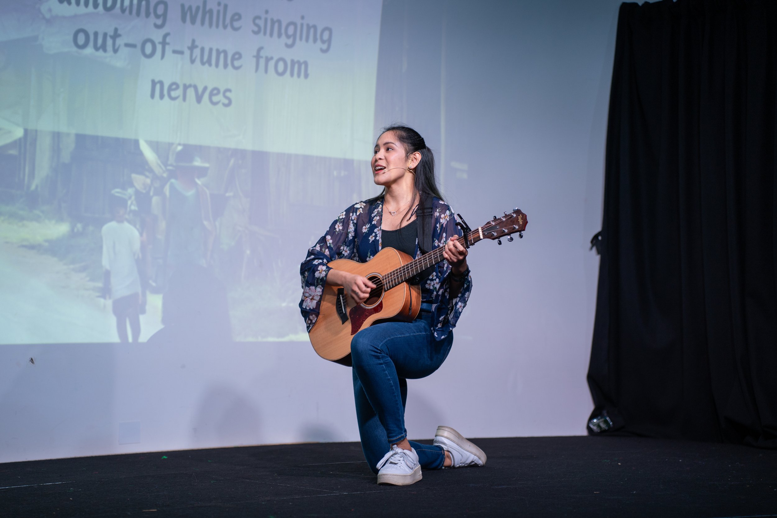A young woman is kneeling on one knee while playing an acoustic guitar on a stage. She is singing and wearing a floral kimono-style jacket, black top, blue jeans, and white sneakers. Behind her is a large screen displaying lyrics and a background image of children and a farm scene.