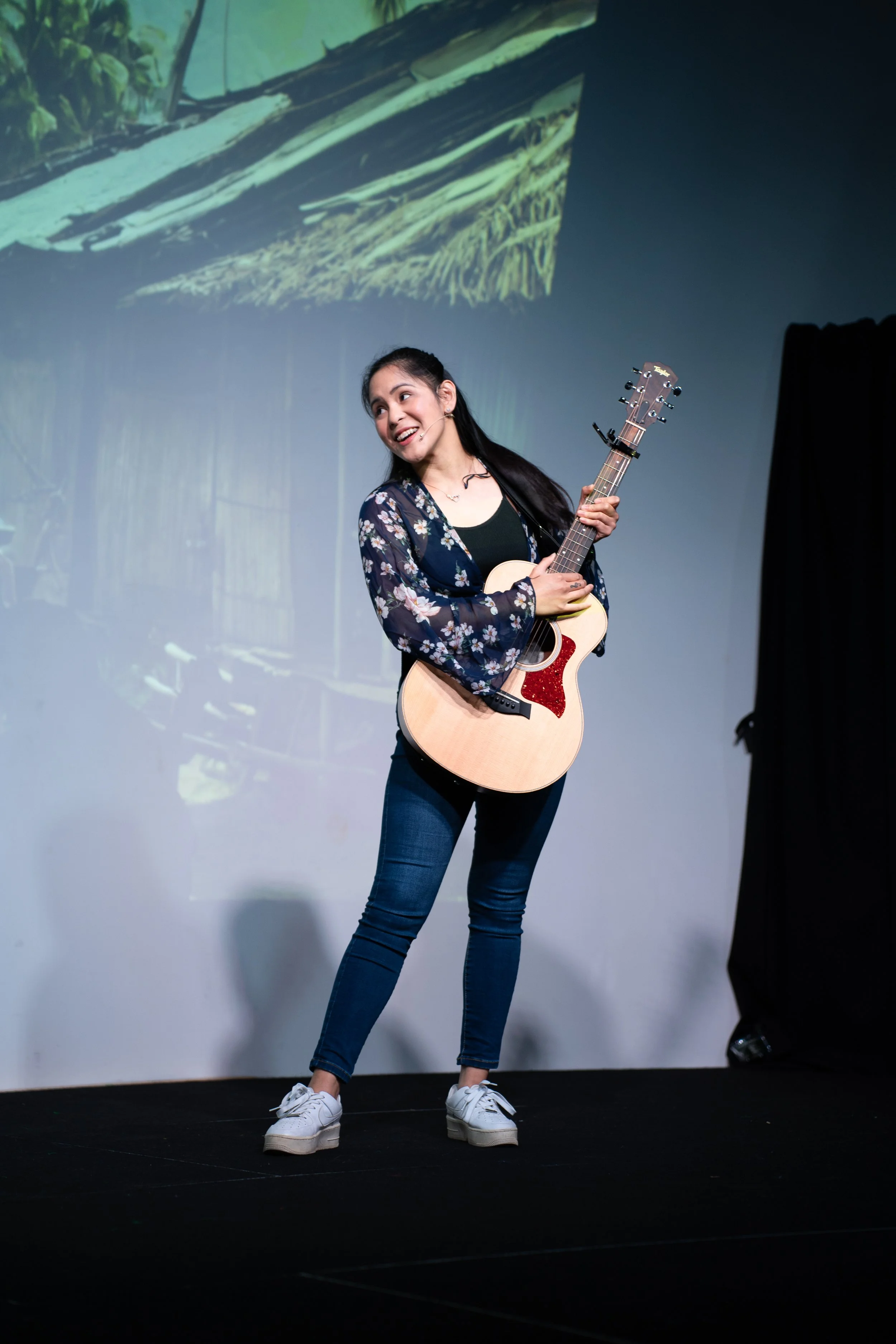 A young woman with long dark hair, wearing a black shirt and floral cardigan, stands on a stage holding an acoustic guitar. She appears to be performing or speaking, with a large screen behind her projecting an image of a thatched-roof hut in a tropical setting.