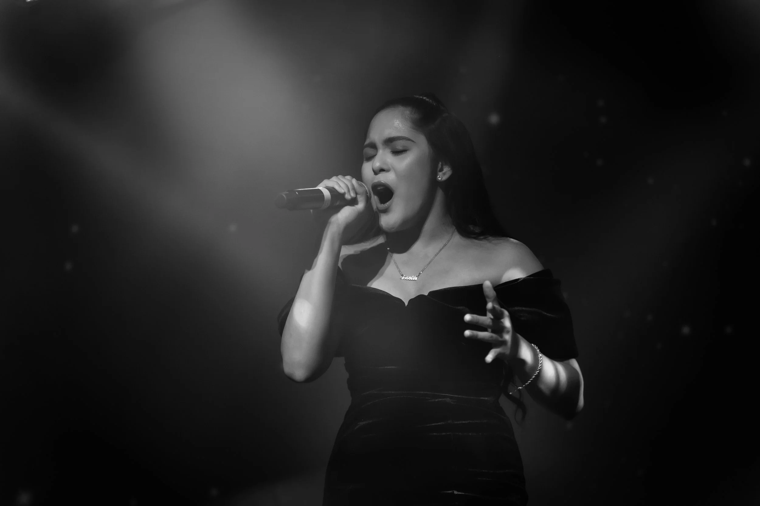 A woman singing passionately into a microphone on stage, with her eyes closed and hand raised, wearing an off-the-shoulder dress and accessories.