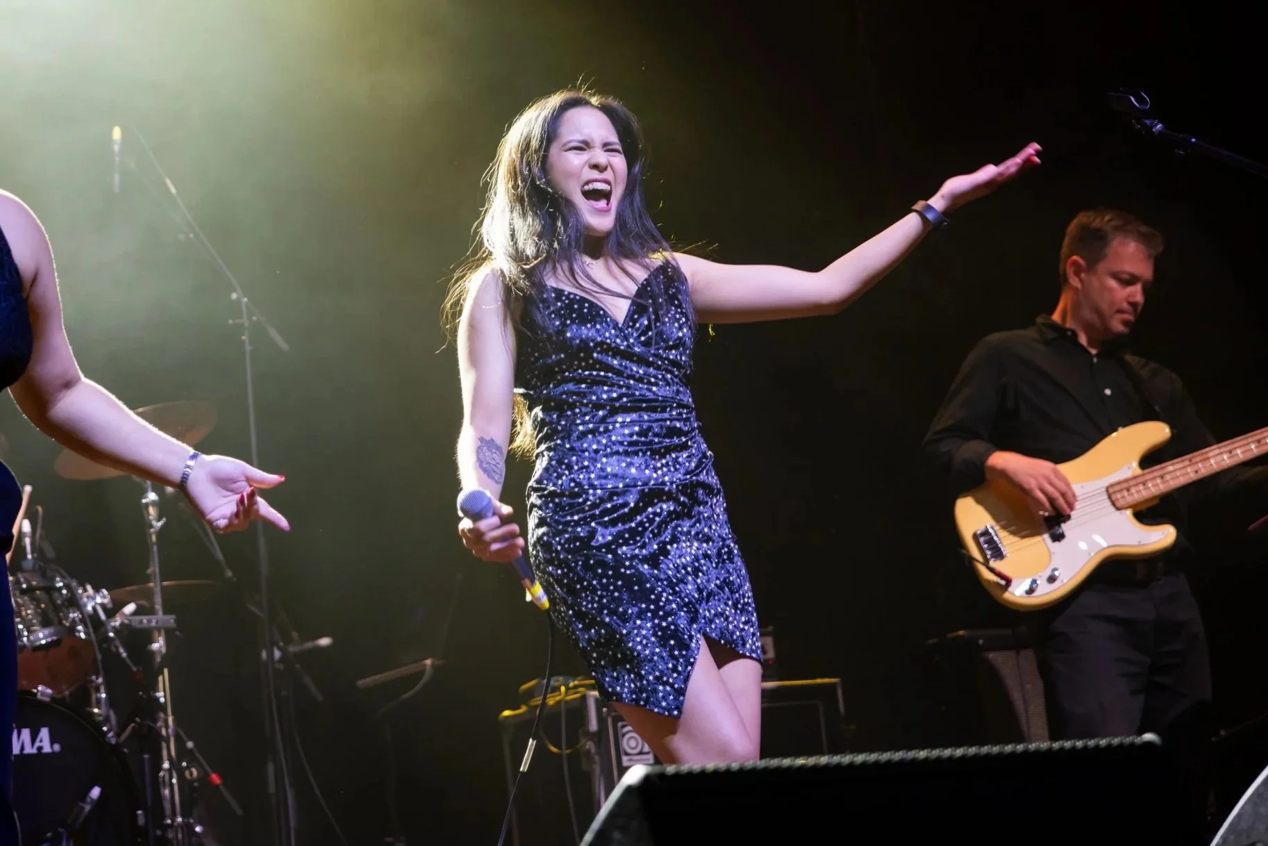 A woman singing on stage, holding a microphone in her right hand, wearing a sparkly dress, with her left arm extended outward. A man is playing a bass guitar beside her.