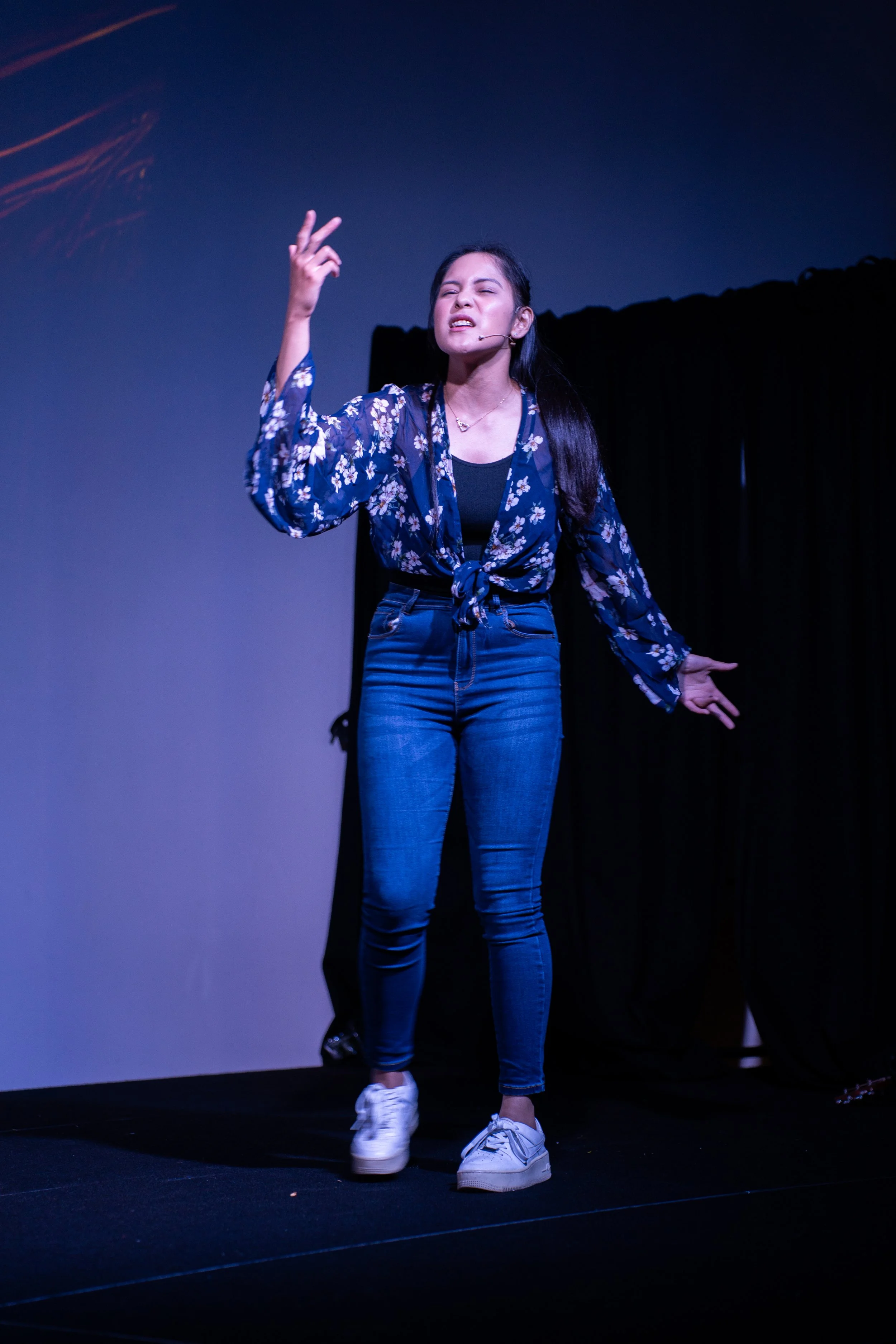 A woman with long dark hair is performing on stage, wearing a blue floral tie-front blouse, blue jeans, and white sneakers. She has a headset microphone and is making expressive hand gestures.