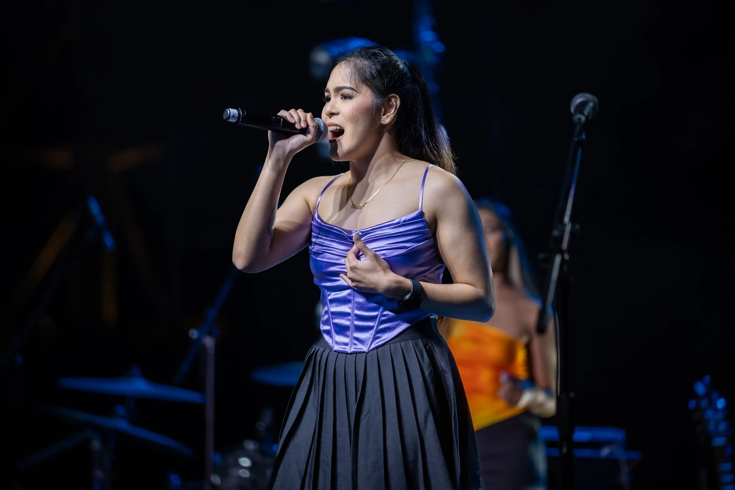 A young woman singing into a microphone on stage, wearing a purple satin top and black pleated skirt, with a wristwatch and a necklace, with a background of musical equipment and another performer in a yellow outfit.