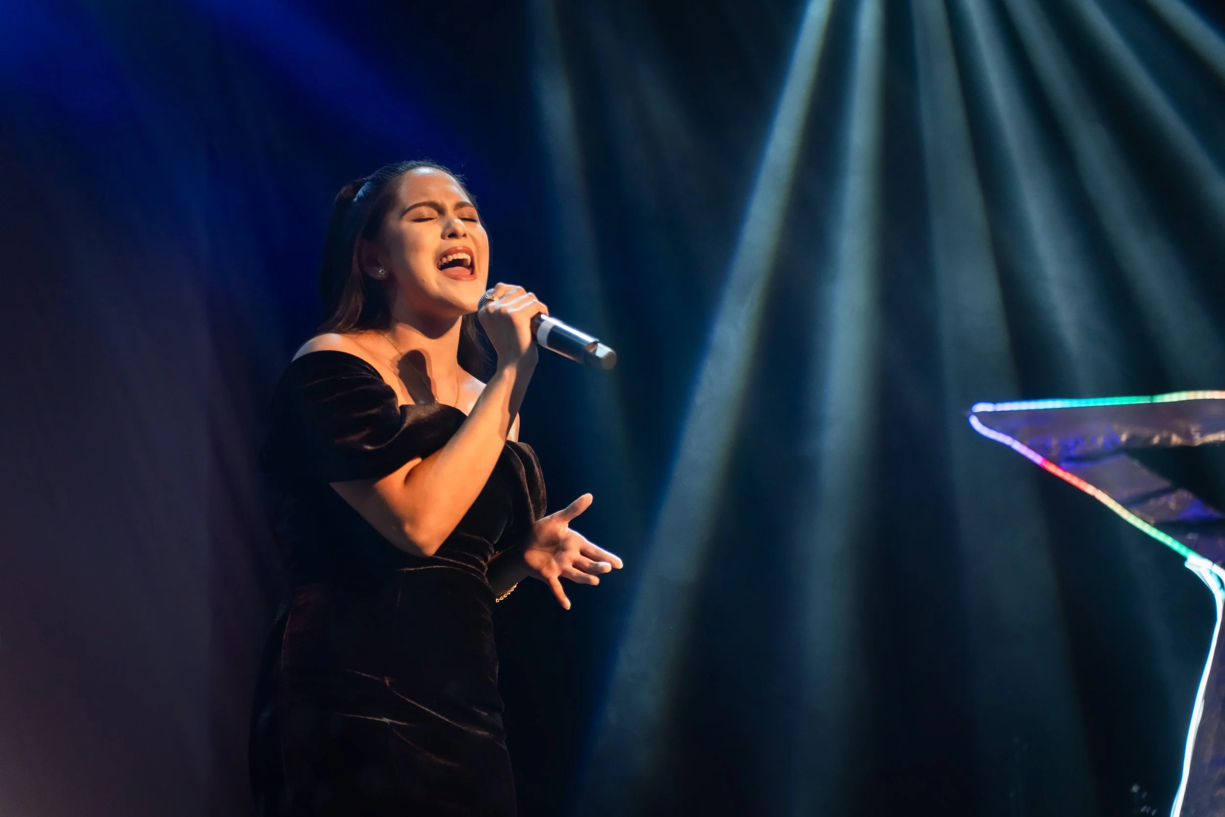 A woman singing passionately on stage with her eyes closed, holding a microphone, illuminated by stage lights, with a colorful grand piano partially visible to her right.