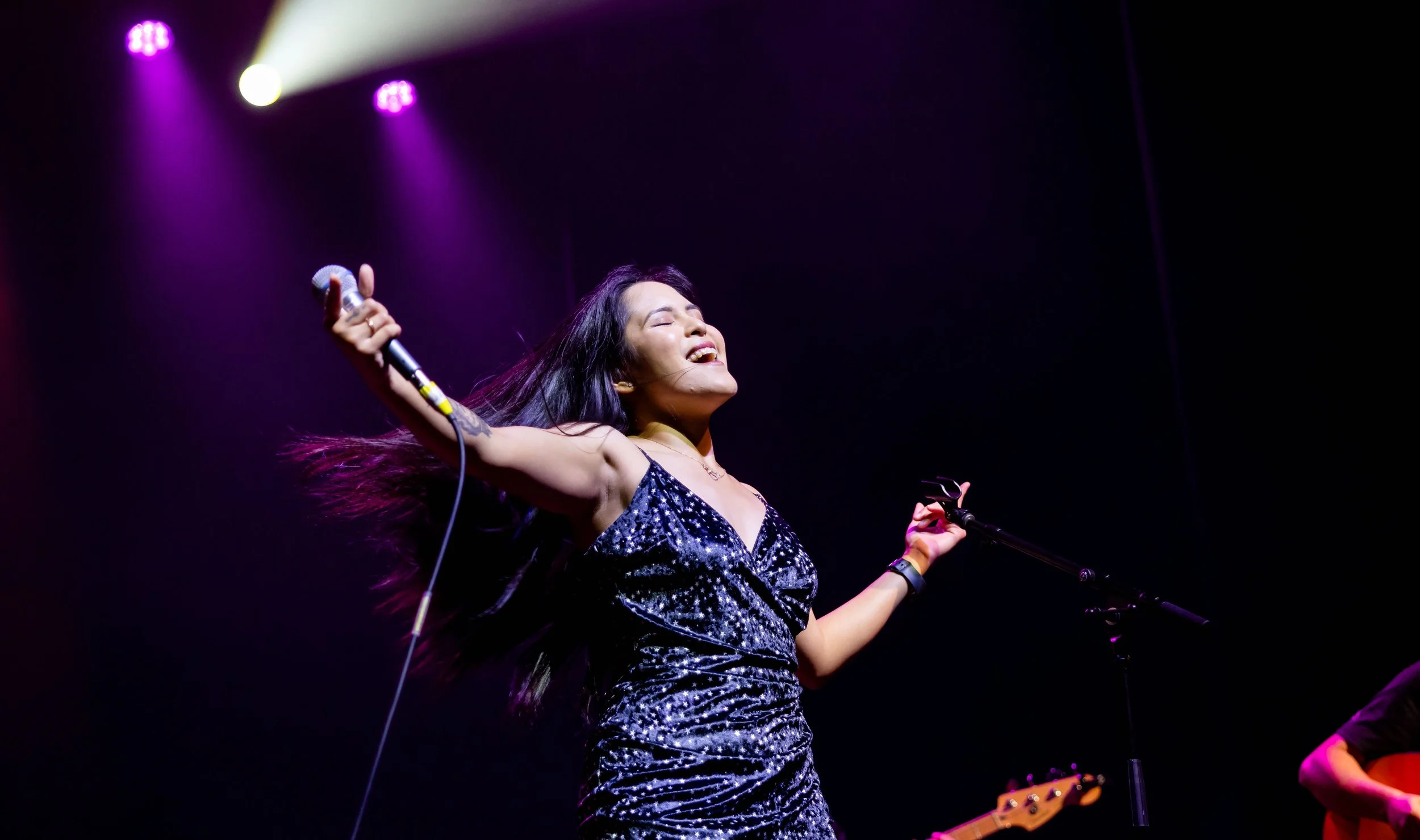 A woman in a sparkling black dress sings on stage with her arms outstretched, eyes closed, under purple stage lights, holding a microphone in her right hand.