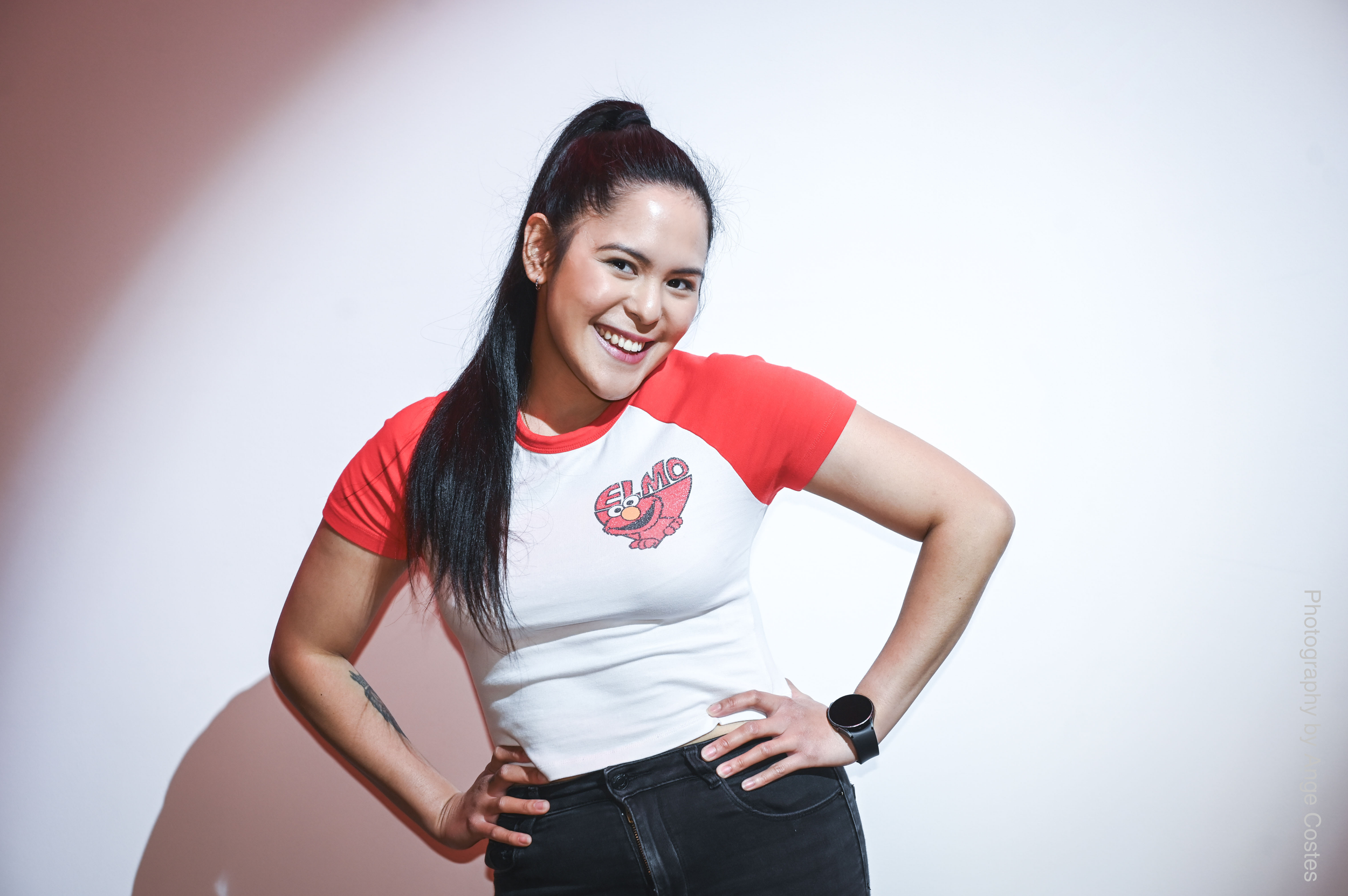 A woman with long black hair, wearing a red and white Elmo t-shirt, black jeans, and a smartwatch, smiling and posing with her hands on her hips against a white background.