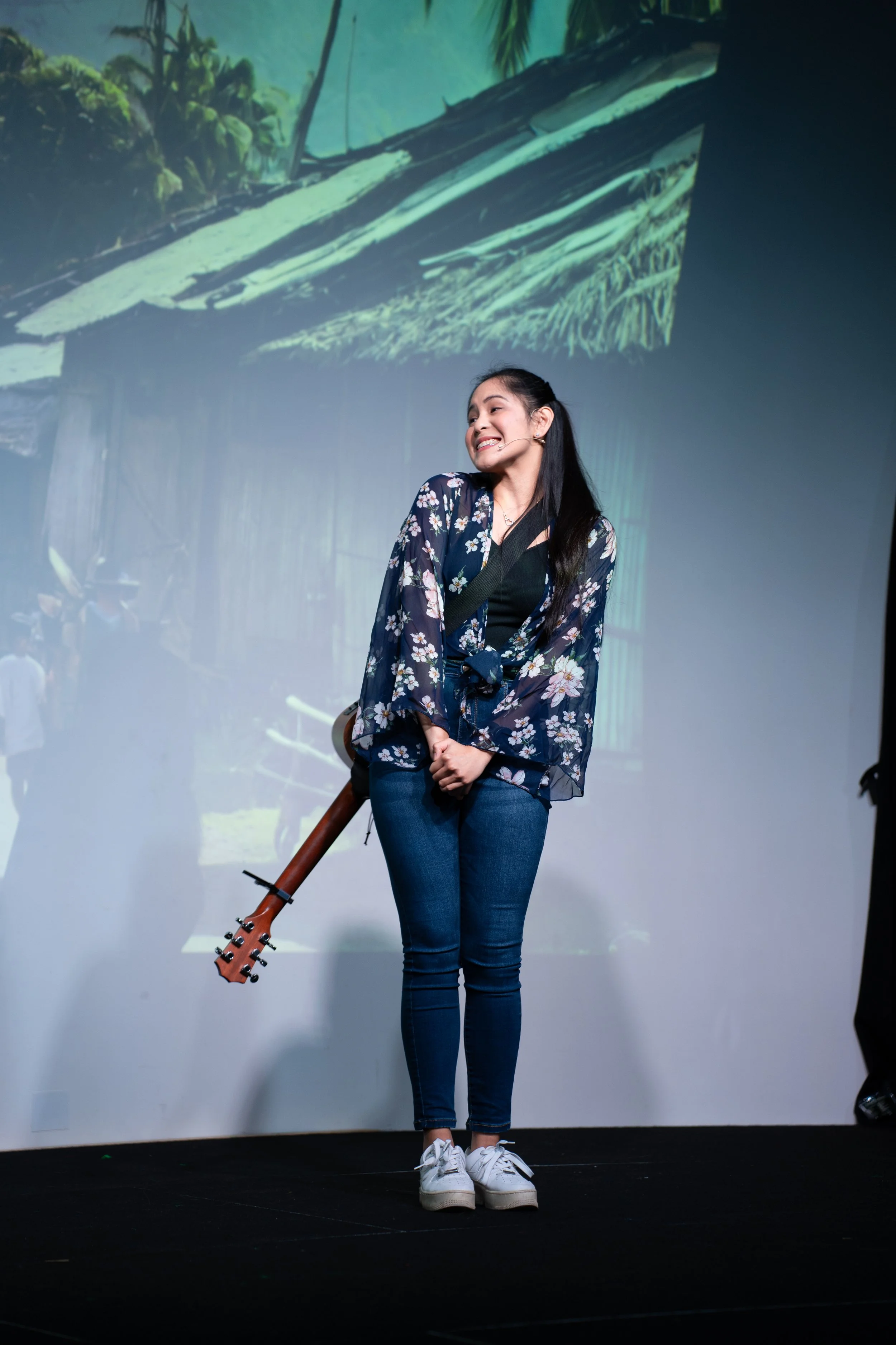 Young woman smiling on stage with a guitar, wearing a floral kimono, jeans, and white sneakers, standing in front of a projection of a rural village scene.