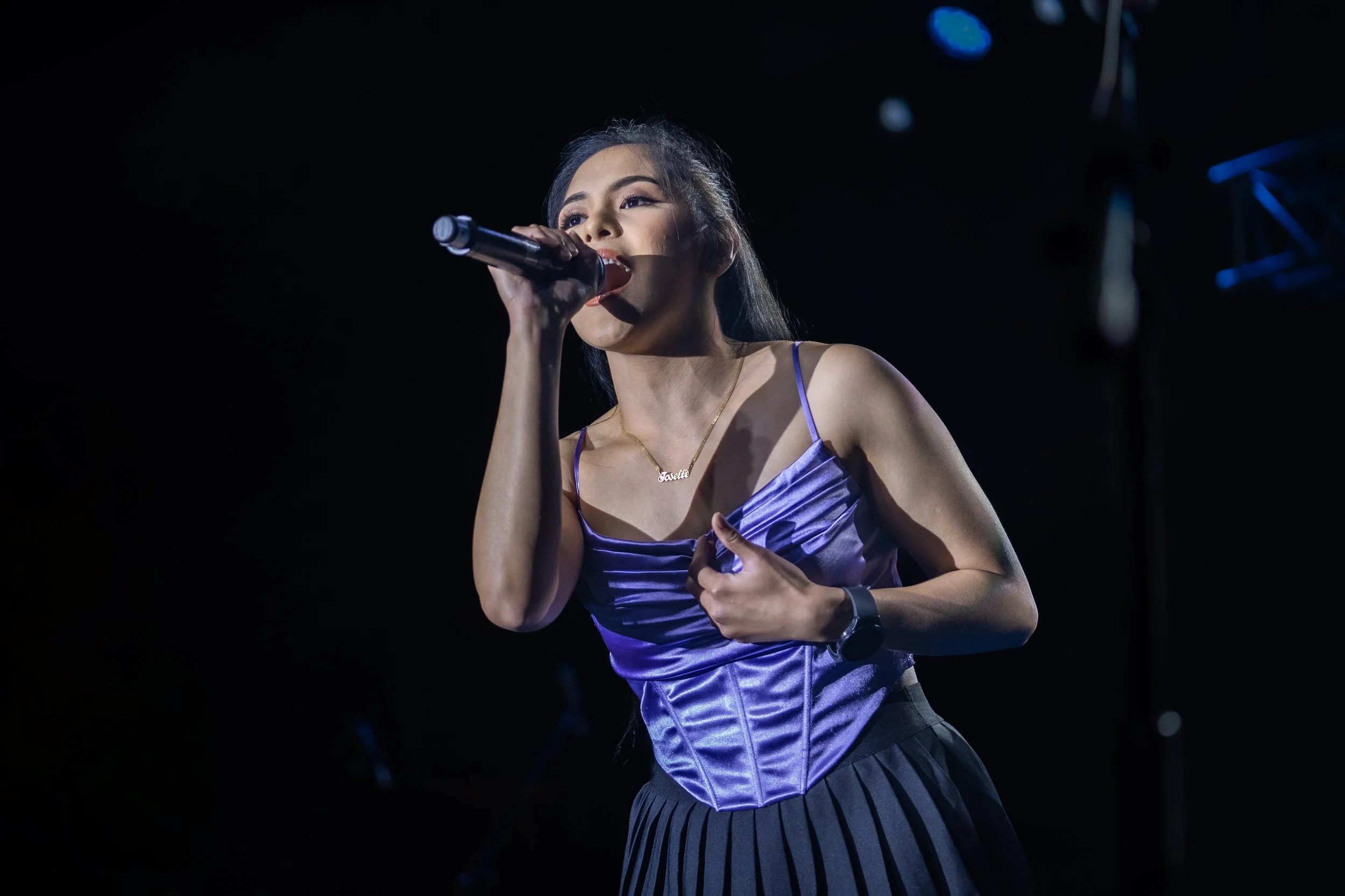 A woman wearing a purple satin top and pleated skirt sings into a microphone on a dark stage.