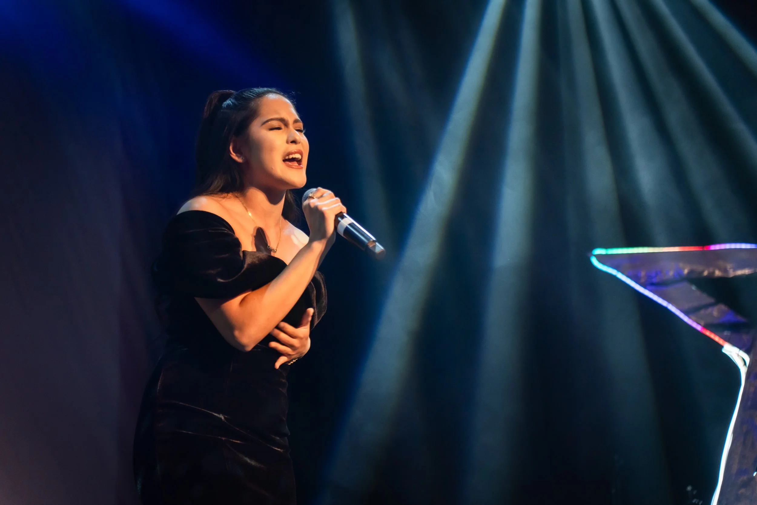 A woman in a black dress singing on stage with a microphone, illuminated by stage lights, with a colorful illuminated grand piano nearby.