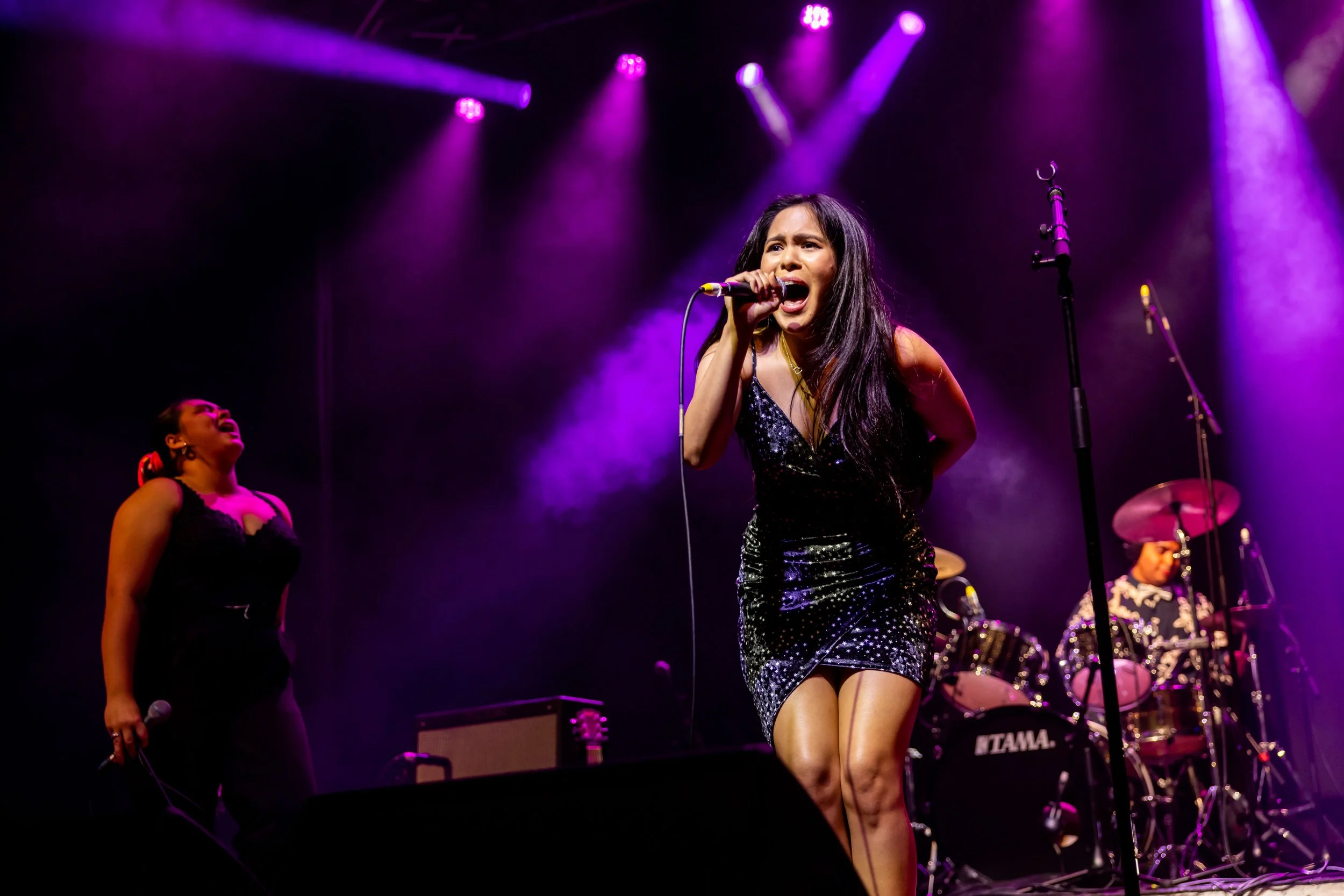A woman singing passionately on stage with purple stage lights, dressed in a black sequined dress. Two backup singers and a drummer are also visible.